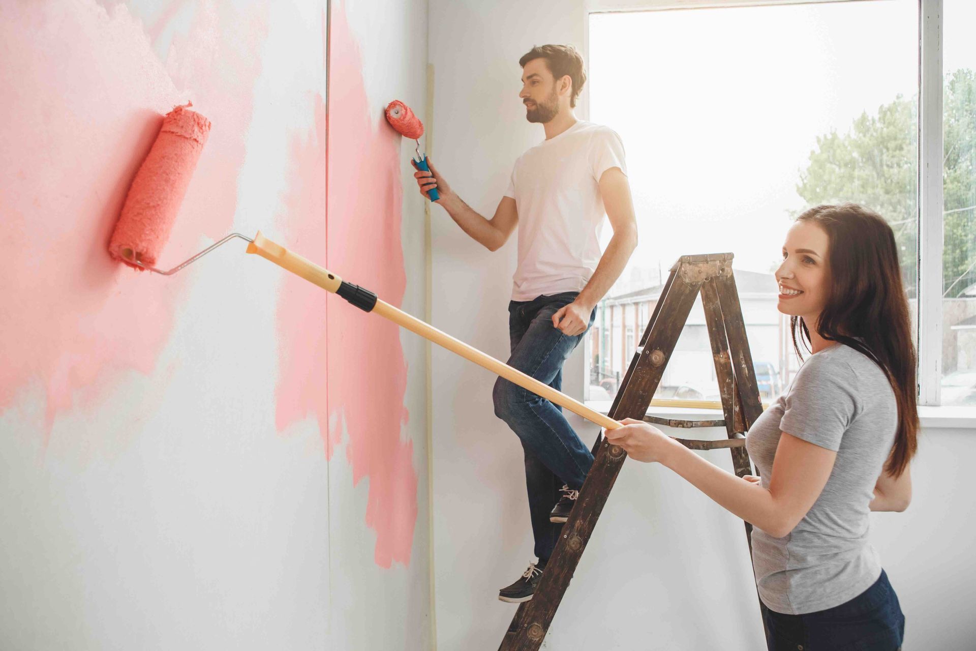 A man is standing on a ladder painting while a woman paints a wall from the ground.