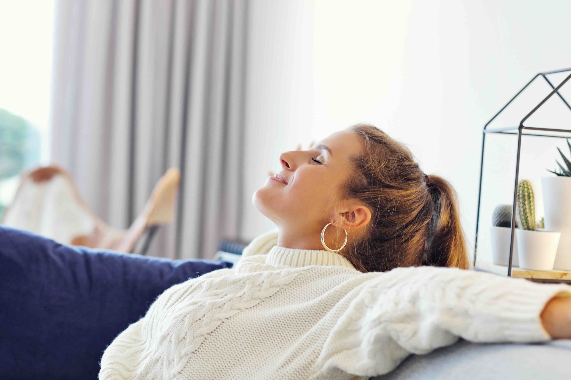 Woman relaxed on a sofa indoors with eyes closed, wearing a white sweater and large hoop earrings.