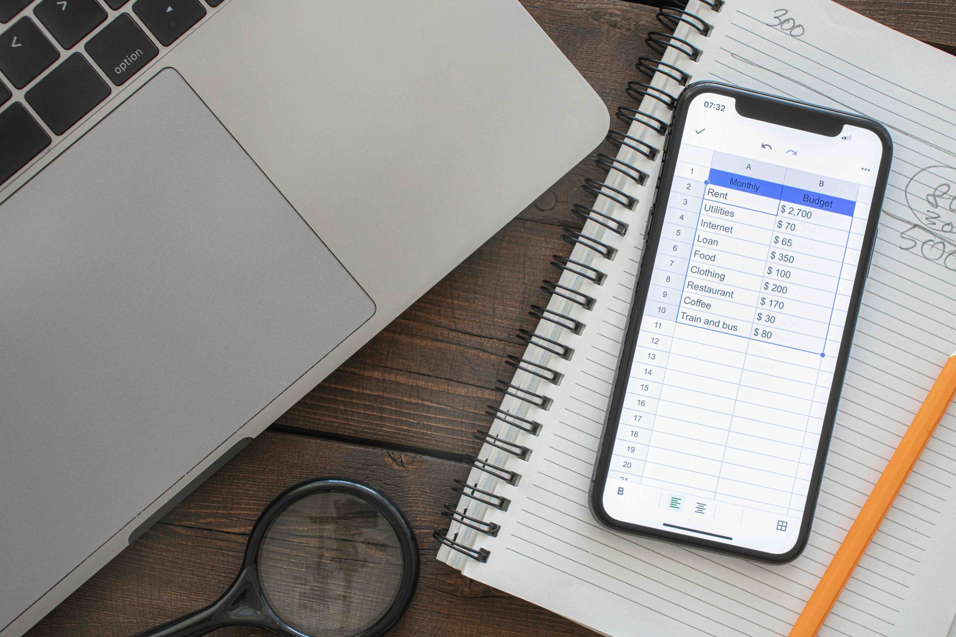 Laptop, smartphone with spreadsheet, notebook, magnifying glass, and pencil on a wooden desk.