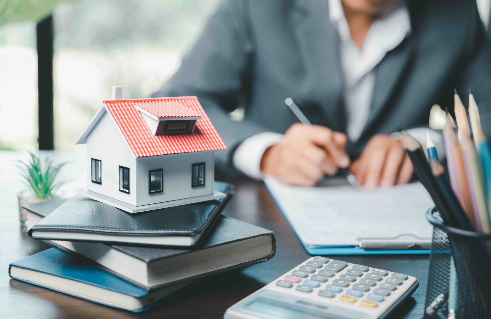 A woman is sitting at a desk with a model house and a calculator figuring out home insurance premiums.