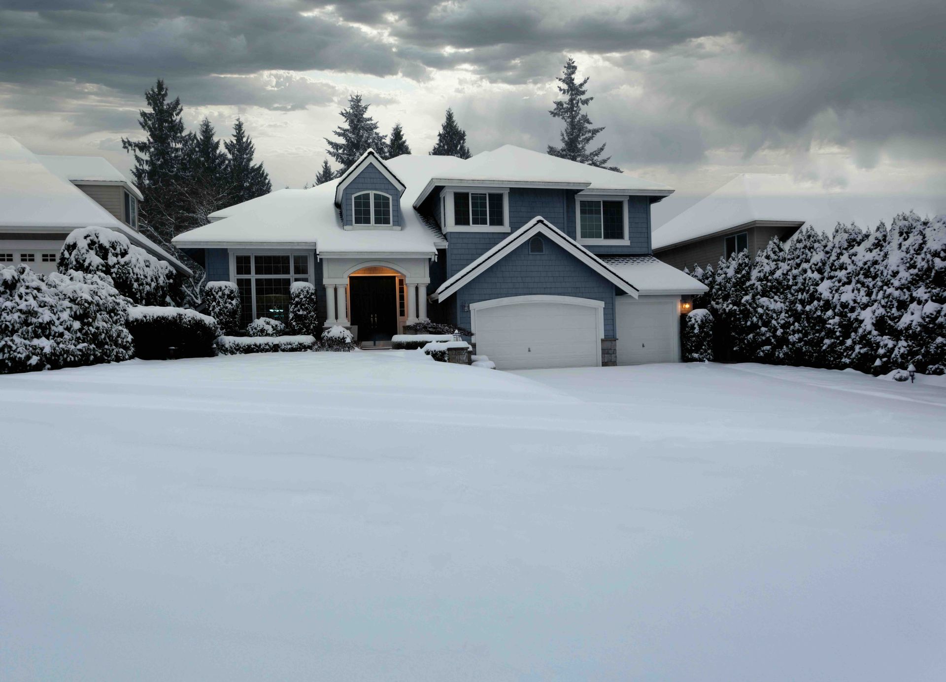 Blue house covered in snow with a cloudy sky. Trees and other houses are also snow-covered.