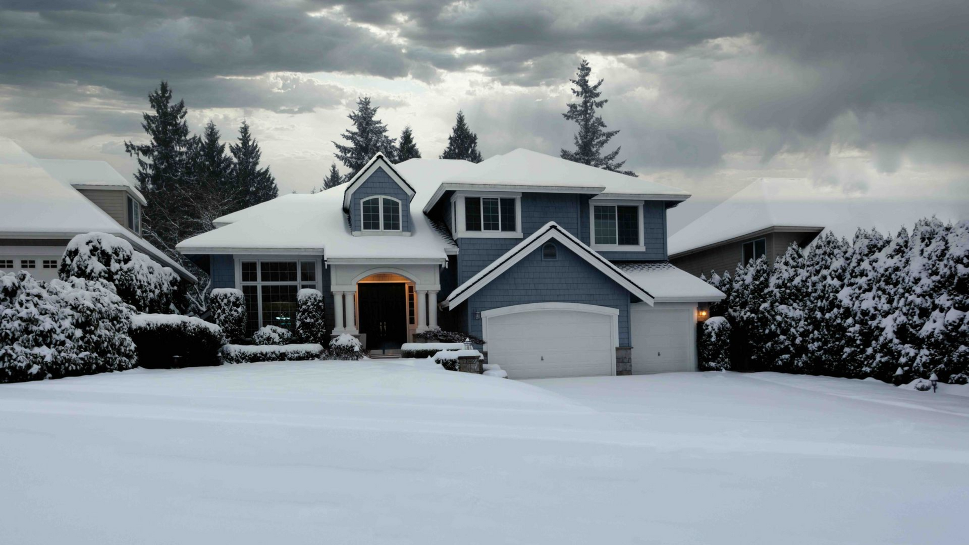 Snow-covered house with blue siding, white garage door, and evergreen trees under a cloudy sky.