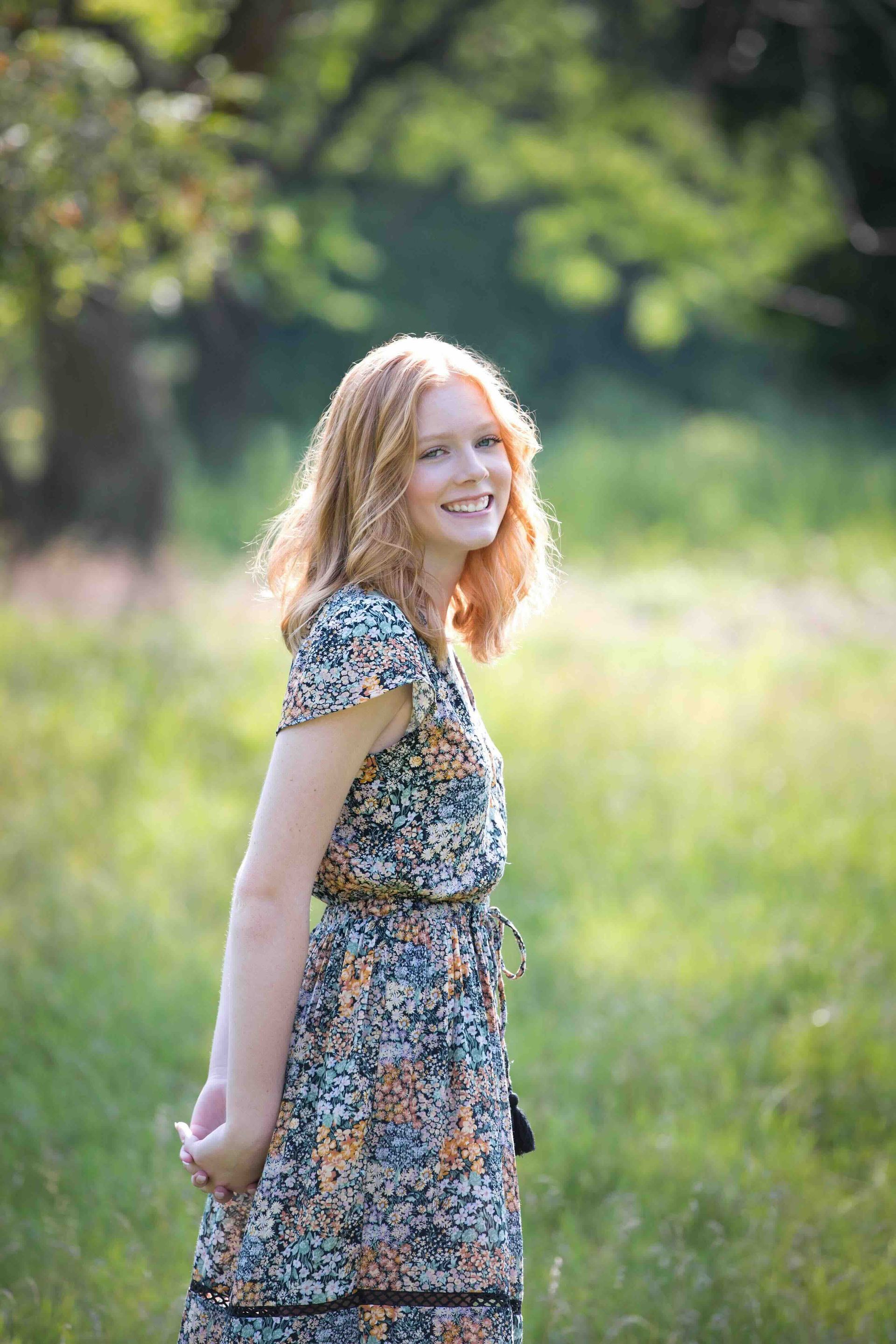 Young person with red hair smiles in a field, wearing a floral dress. Green background.