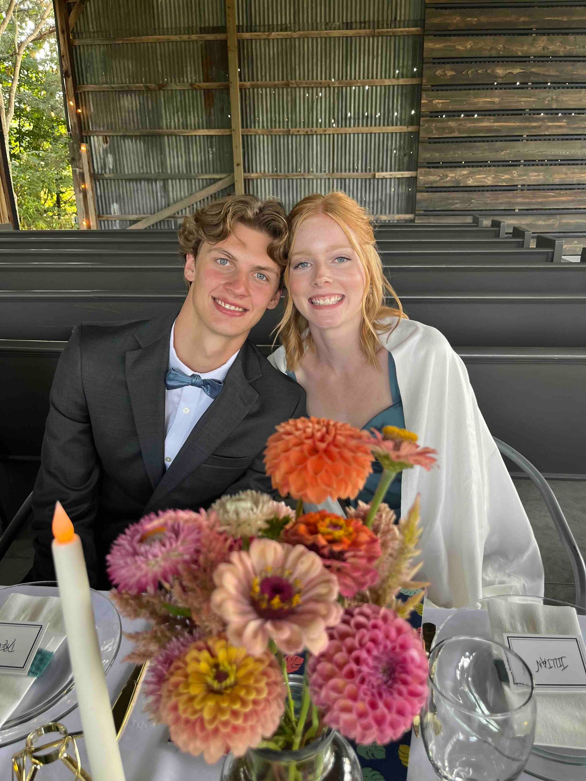 Couple smiling at a table with flowers. The man wears a suit with a bow tie, and the woman has a shawl.