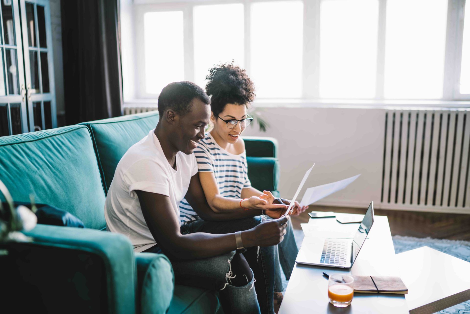 Couple on teal couch reviewing papers, with laptop on table and window in background.