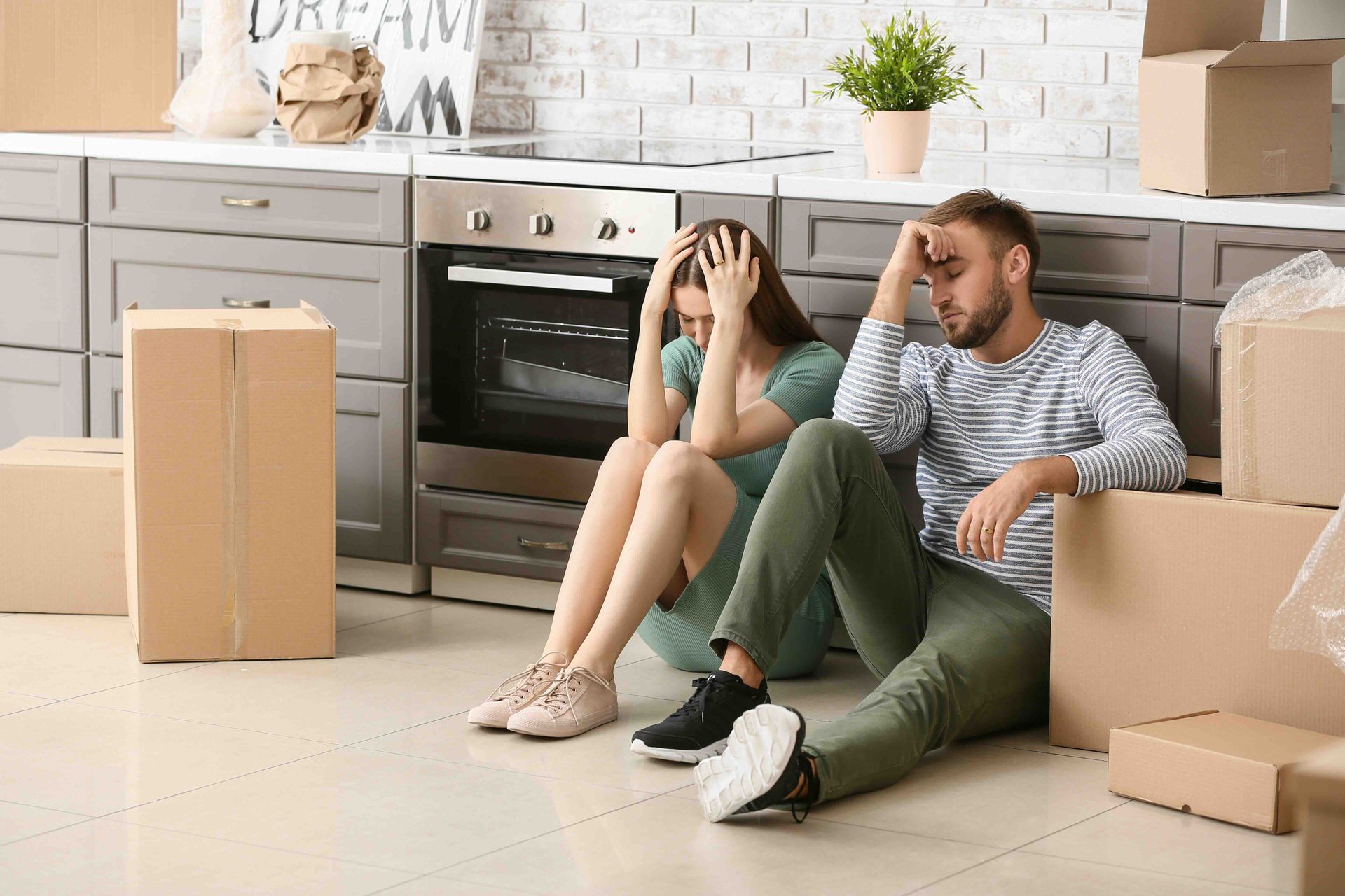 A man and a woman are sitting on the floor in a kitchen surrounded by cardboard boxes.