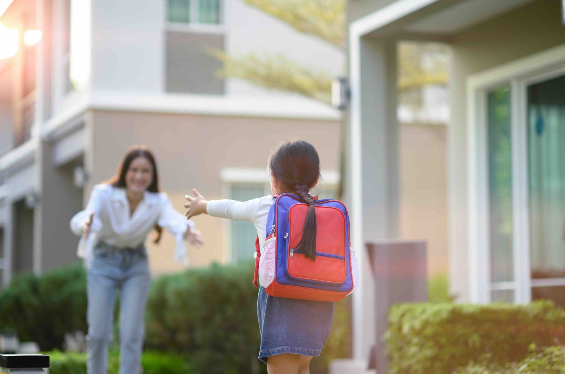 A child with a backpack reaches toward an adult with open arms outside a house.