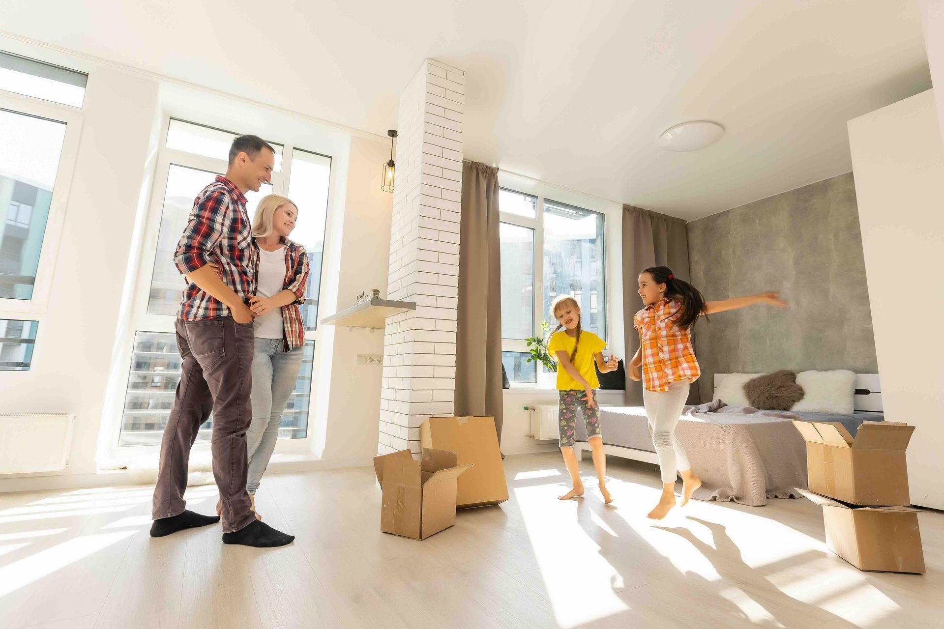 A family moves into a new apartment, with parents watching as two children jump happily near unpacked cardboard boxes.