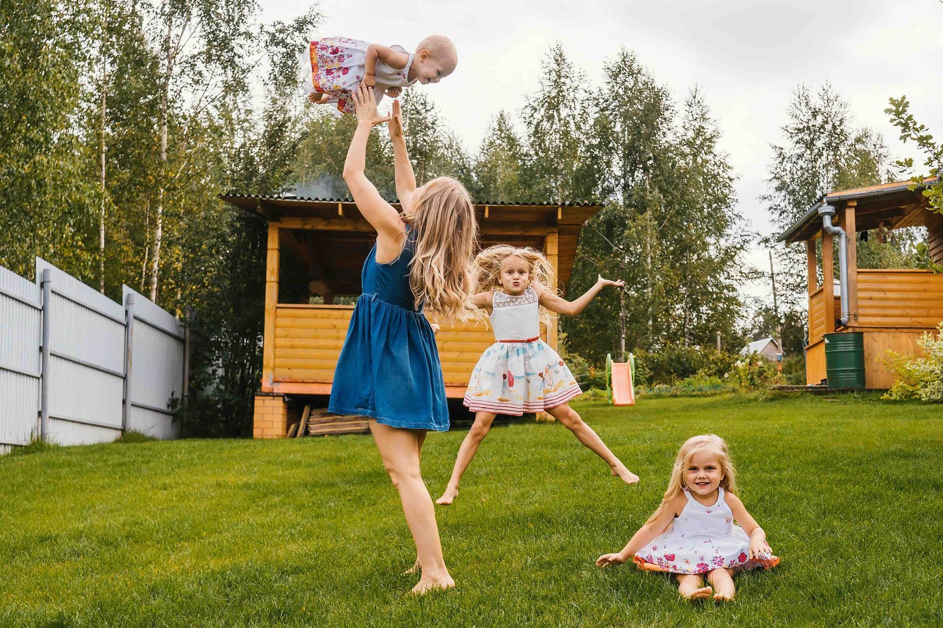 Three little girls are playing in the grass in a backyard with their mother.