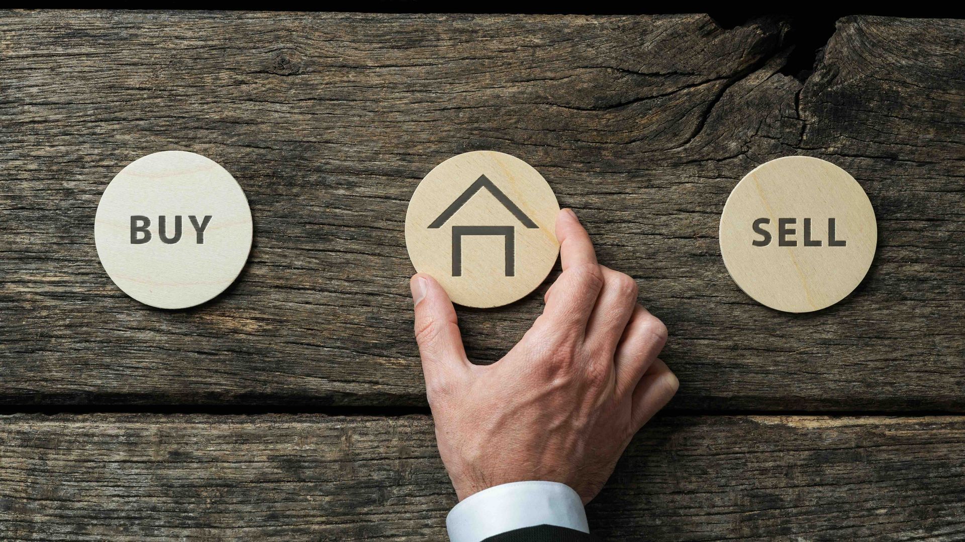 a person is holding a wooden circle with a house on it with another circle on the left that says buy and one on the right that says sell.