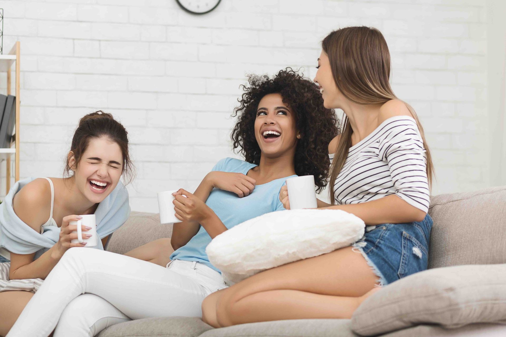 Three people laughing on a couch, holding mugs, in a living room.