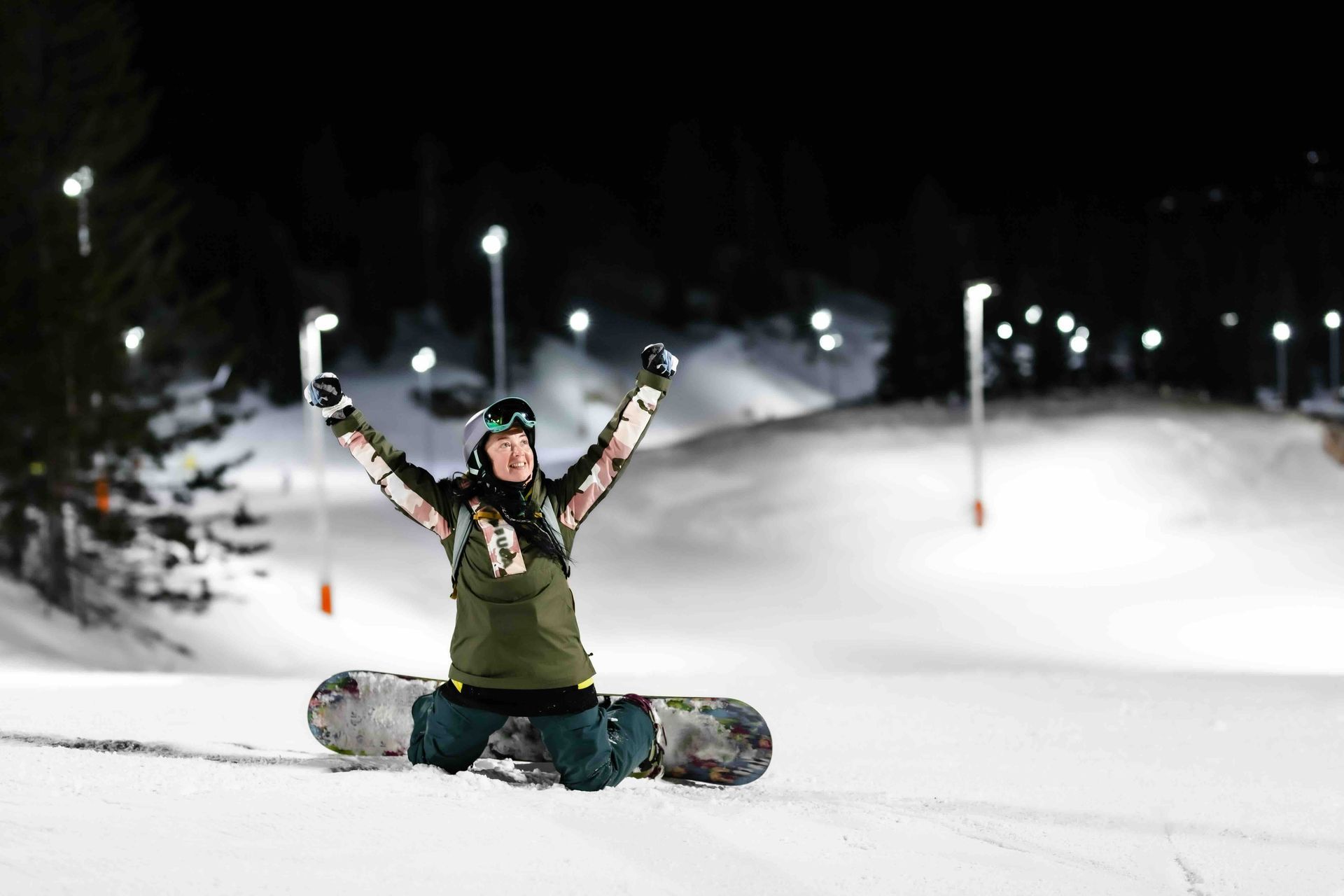 A person is kneeling on a snowboard with their arms in the air.