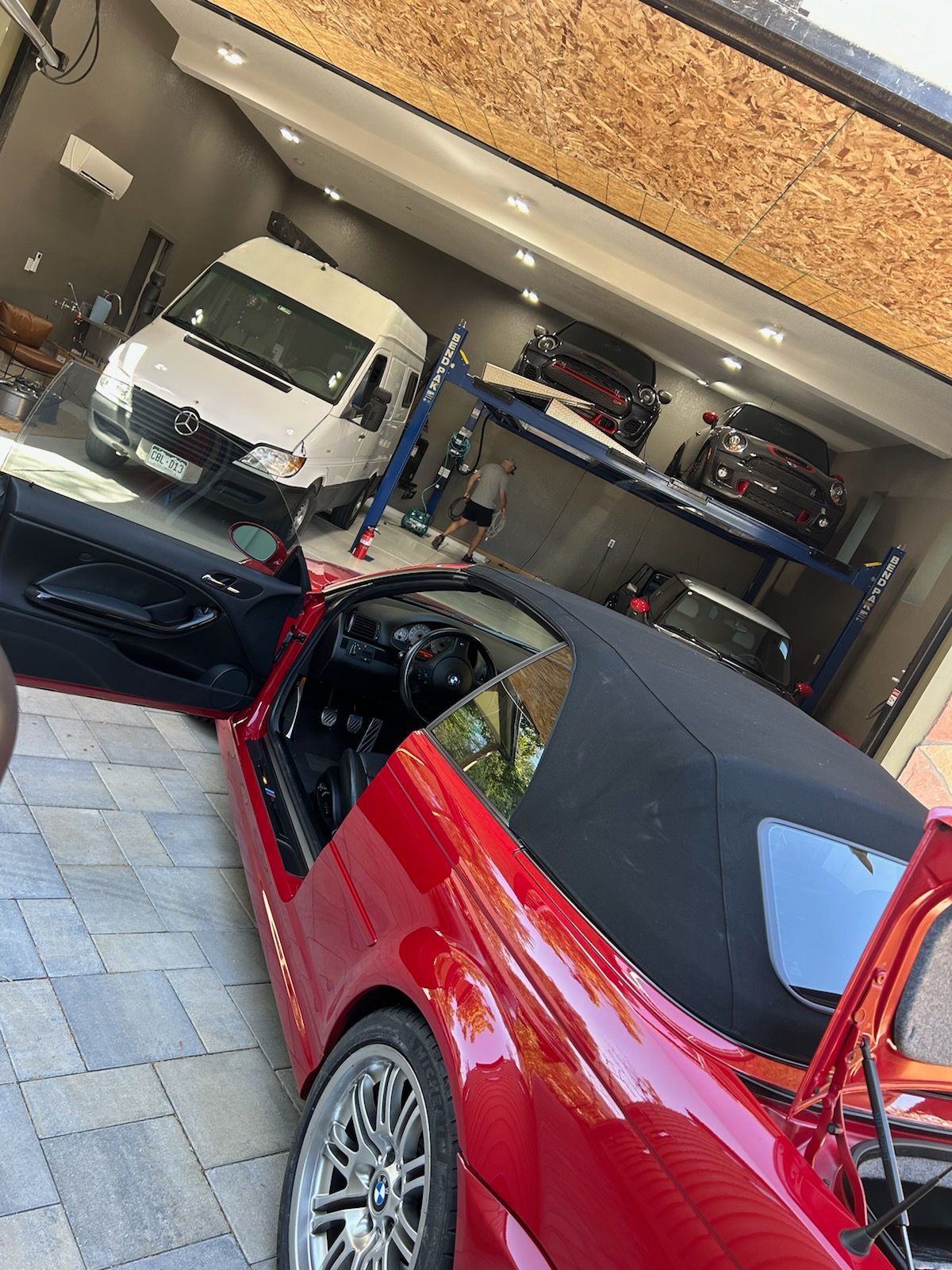 Red convertible in garage, door and trunk open. White van, black cars on a lift. Brick paved floor.