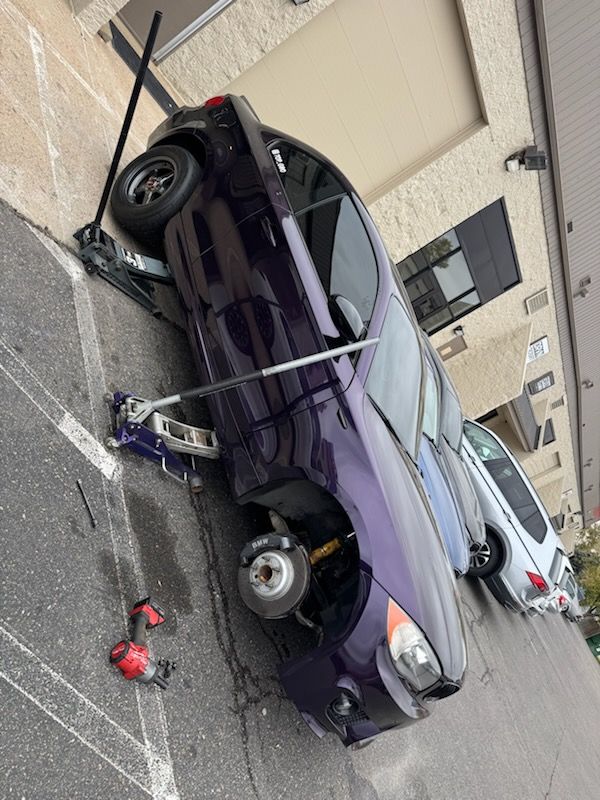 Purple car raised on jacks in a parking lot, one wheel removed, with tools nearby.