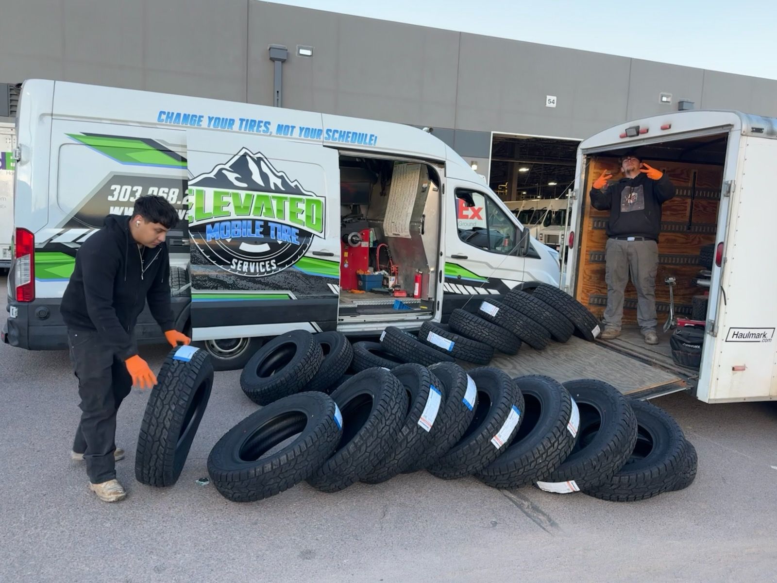 Two men loading tires from a van and trailer outside. 