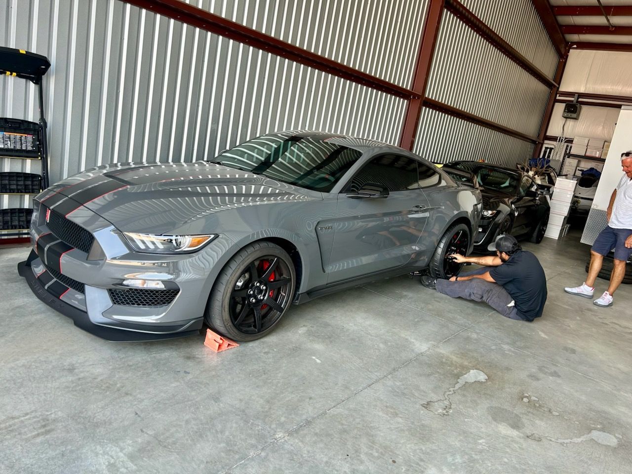 Gray Ford Mustang Shelby GT350 being worked on in a garage by a person. Another person watches.