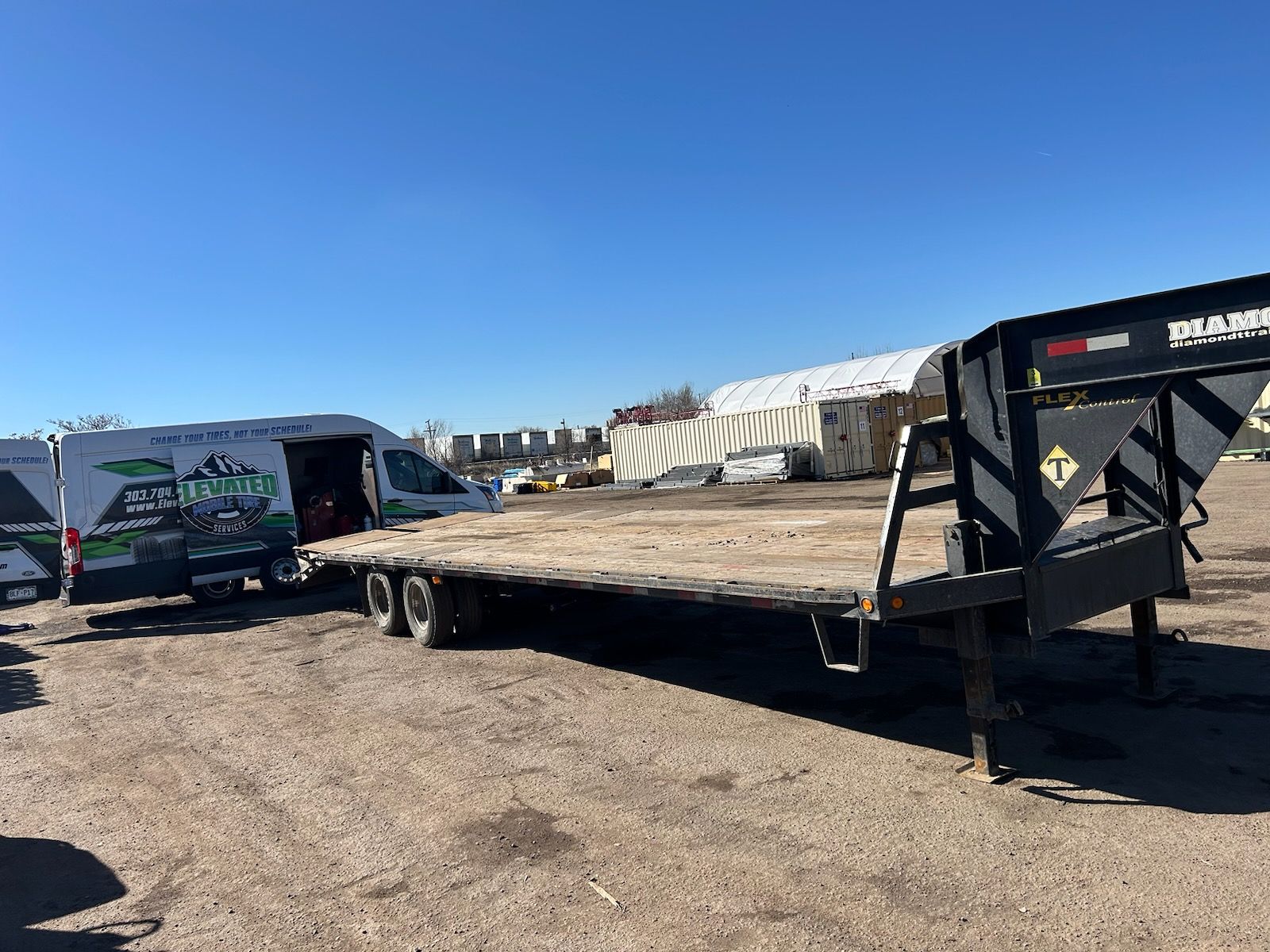Flatbed trailer in a gravel lot next to a van with a company logo under a clear blue sky.
