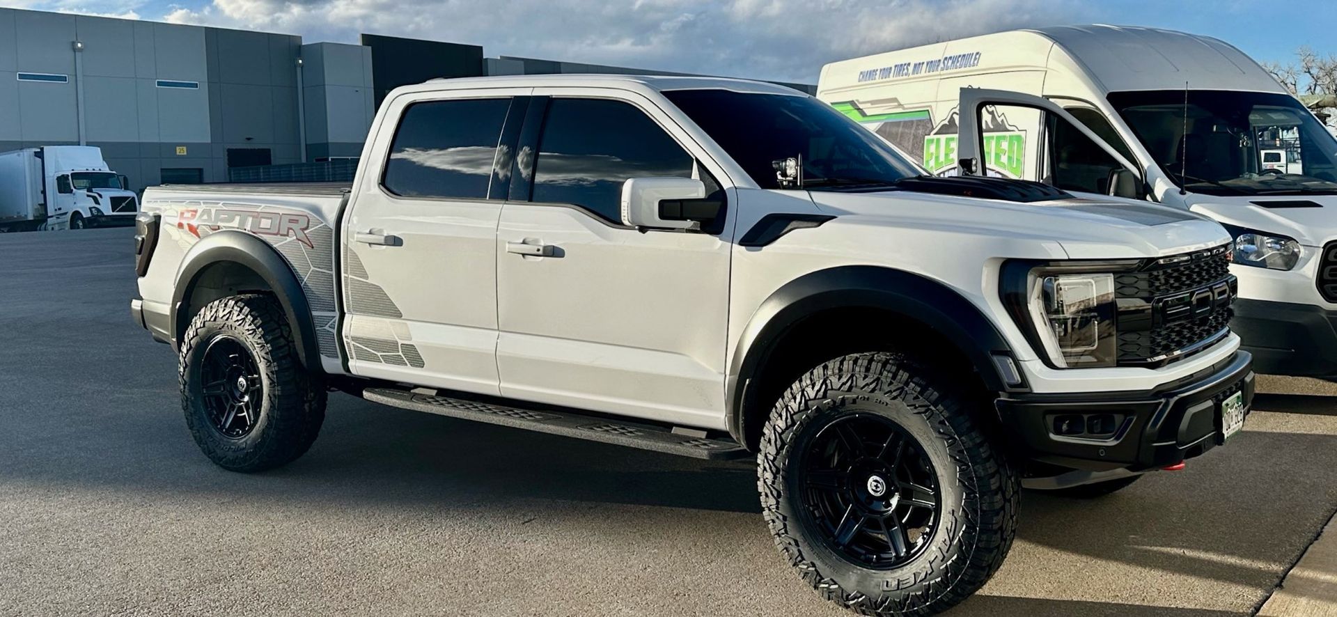 White Ford Raptor truck with black wheels parked on pavement. Van in background.