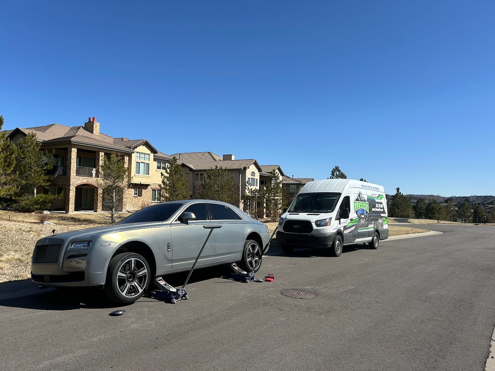 Silver Rolls-Royce with a flat tire, being serviced by a white service van, on a suburban street.