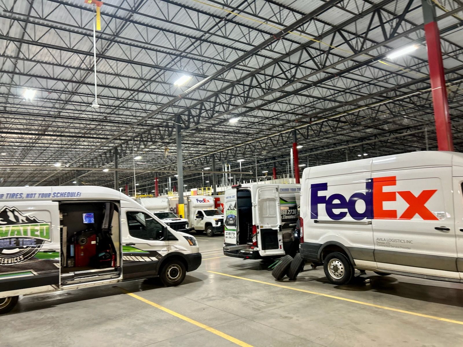 FedEx and delivery vans parked inside a large warehouse with high ceilings and metal beams.