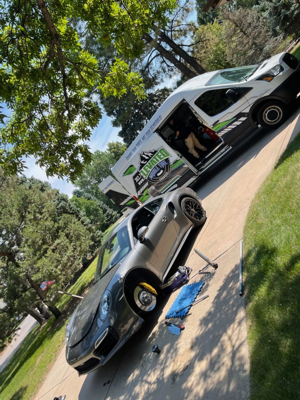 Gray sports car being serviced next to a white van on a driveway. Green grass and trees in the background.
