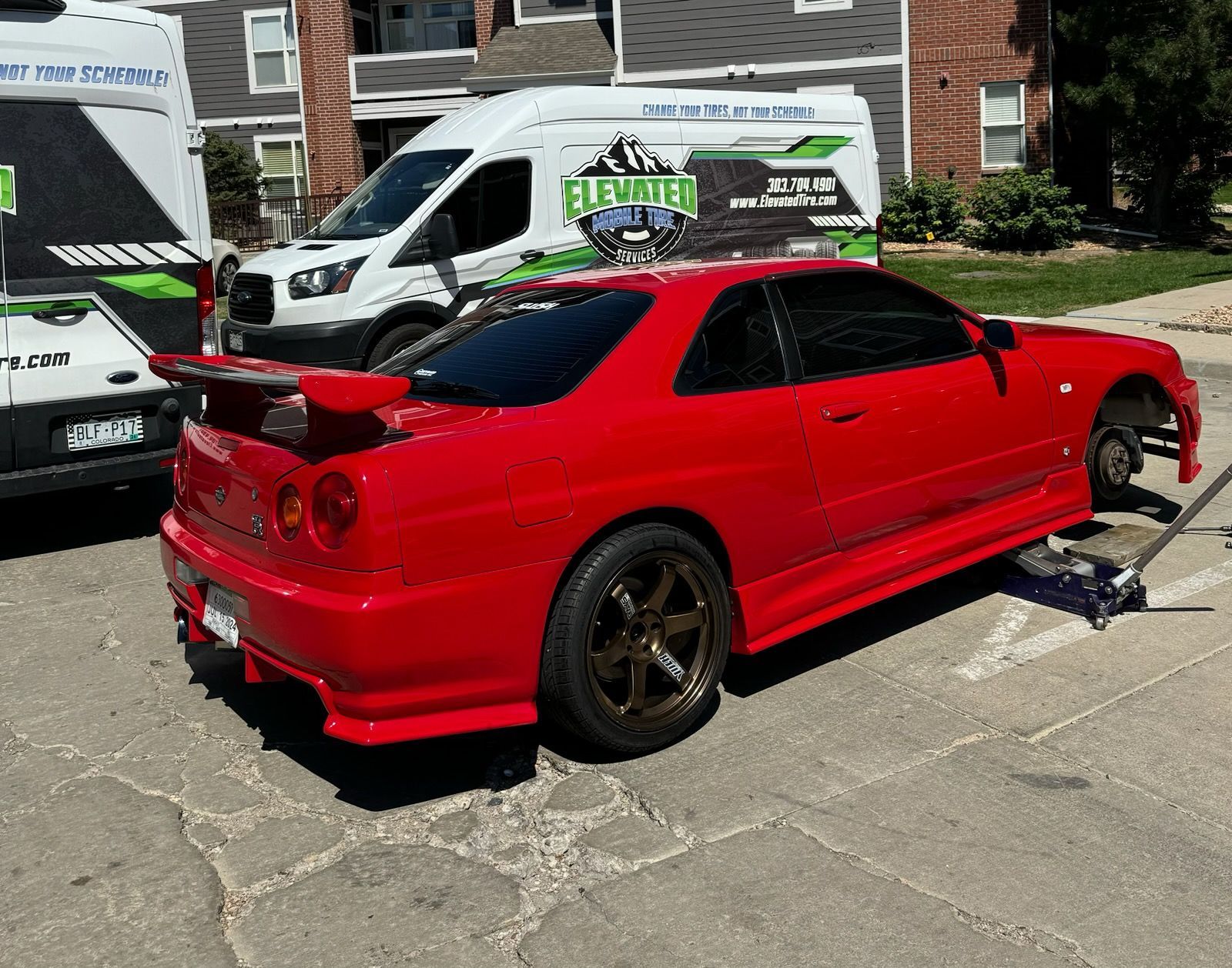 Red sports car with a missing wheel being worked on outdoors, near service vans.
