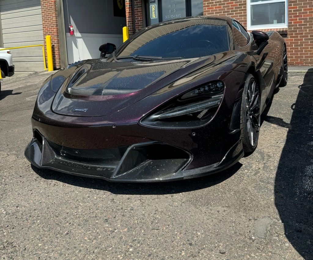 A dark purple McLaren 720S sports car parked in front of a brick building.