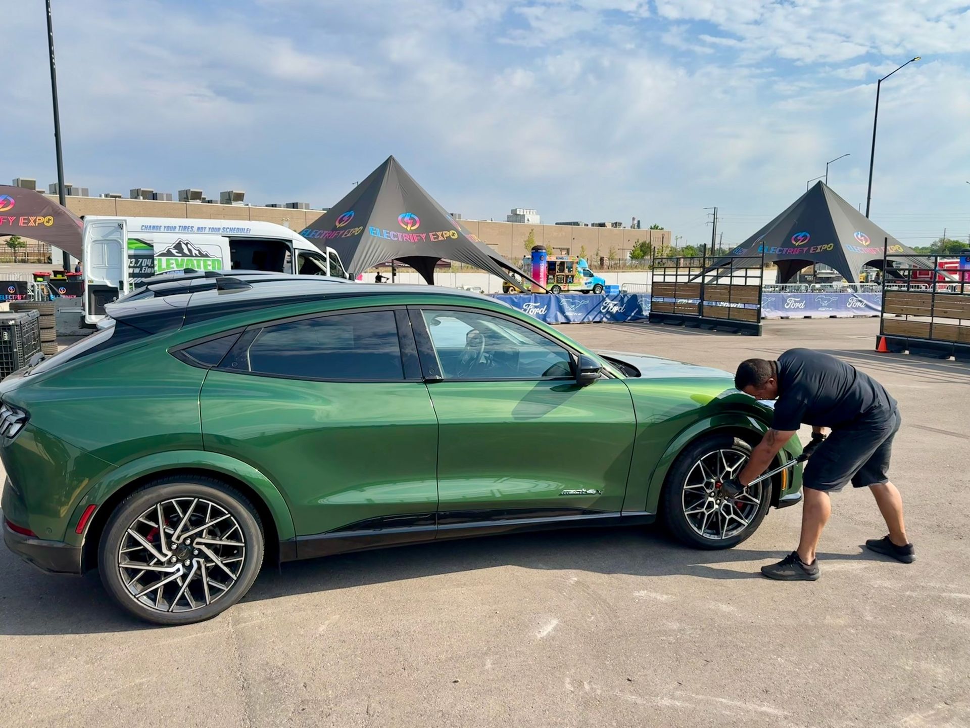 Green Ford Mustang Mach-E car parked on asphalt with a man working on the front wheel, outdoor setting.