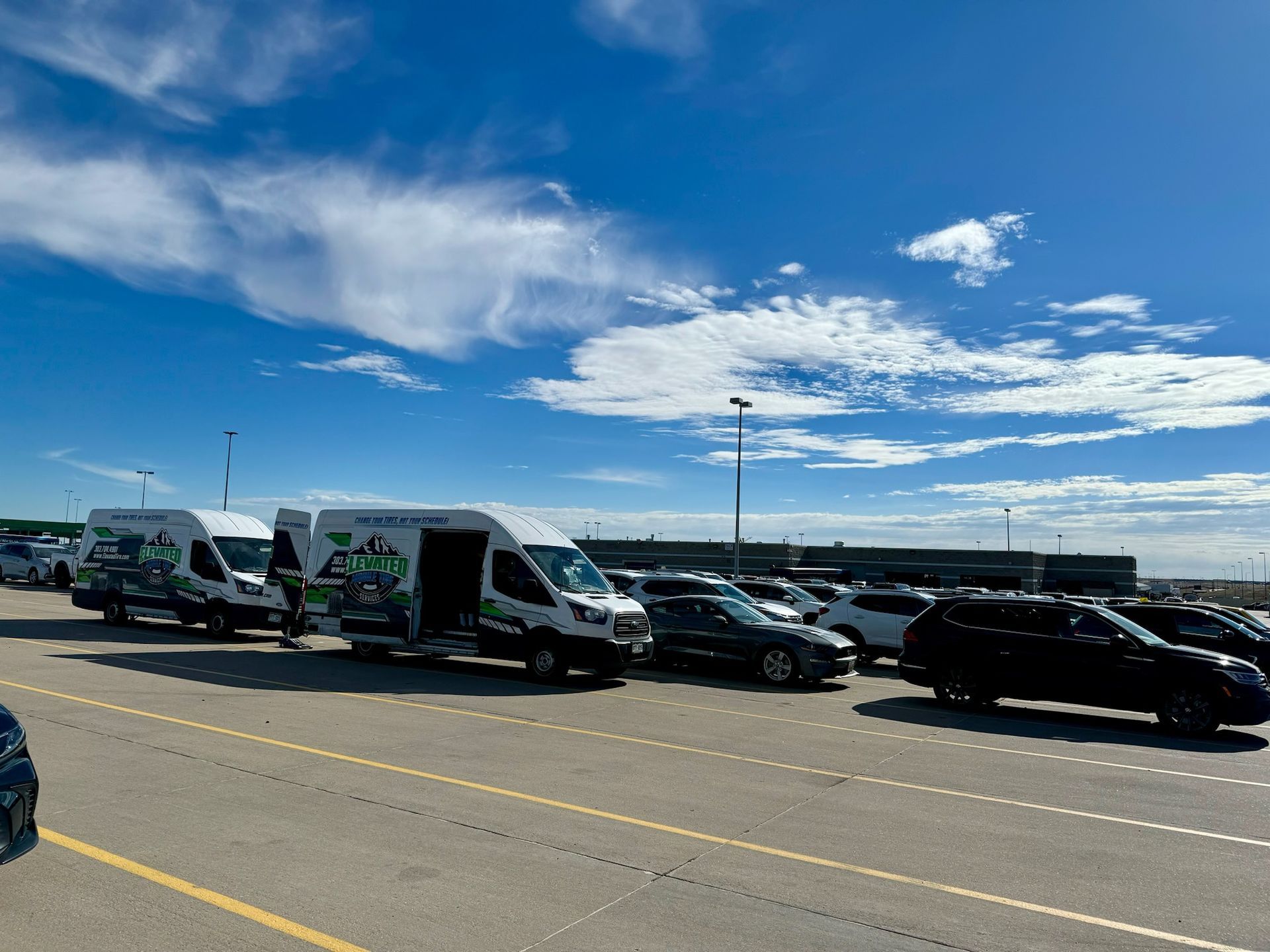 Several white vans with green logos parked in a lot with cars under a blue sky with clouds.