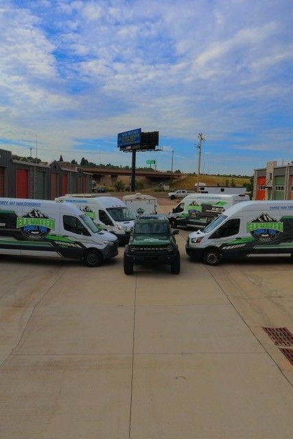 A dark green Jeep surrounded by white vans with green logos in a parking lot under a cloudy sky.