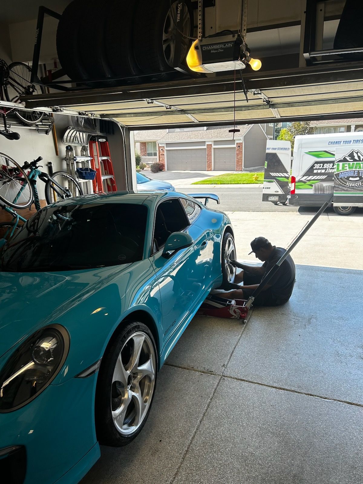 A mechanic changing a tire on a bright blue Porsche in a garage; a service van is visible outside.