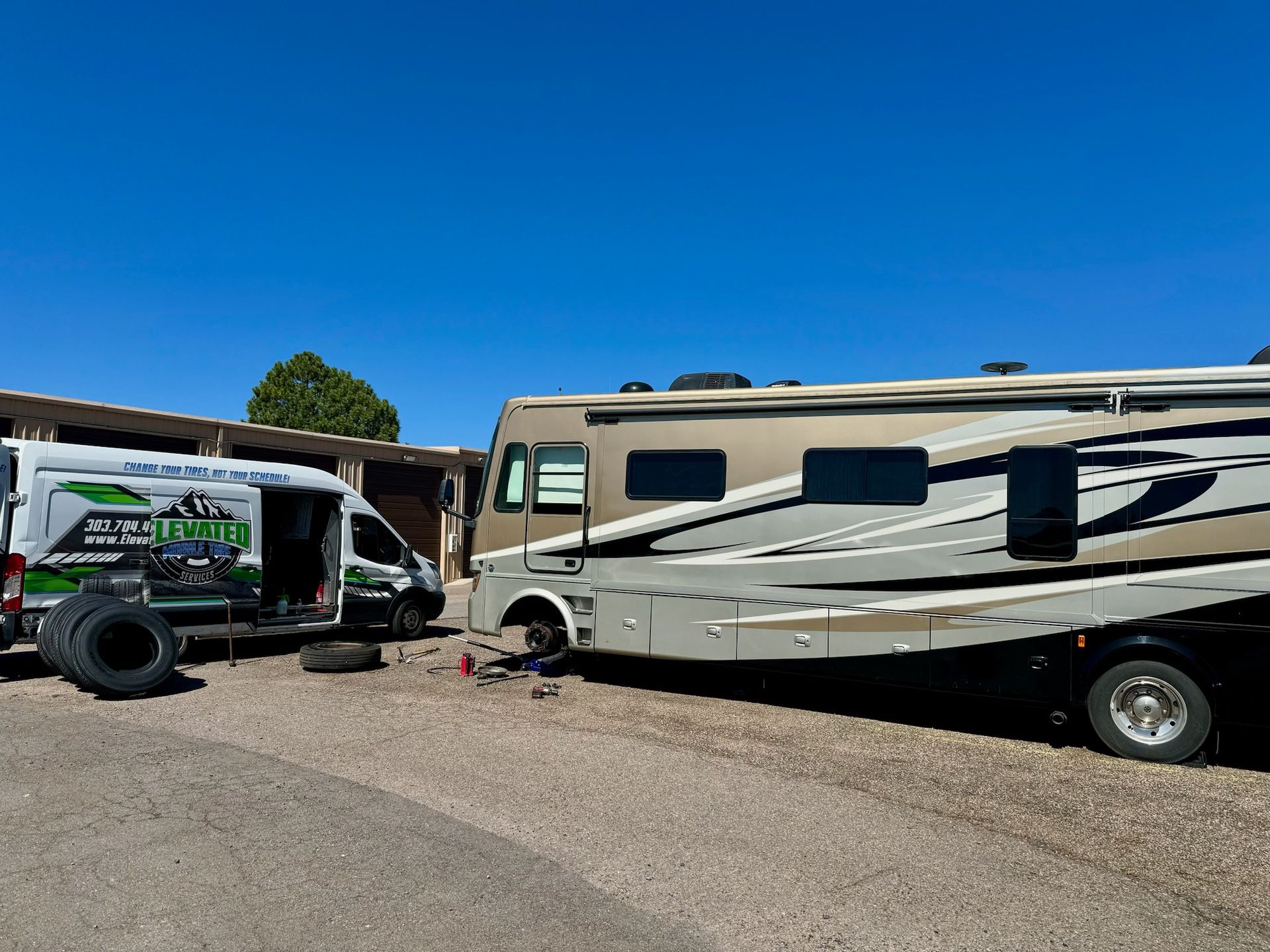 A motorhome being serviced by a mobile tire repair van on a gravel lot under a clear blue sky.