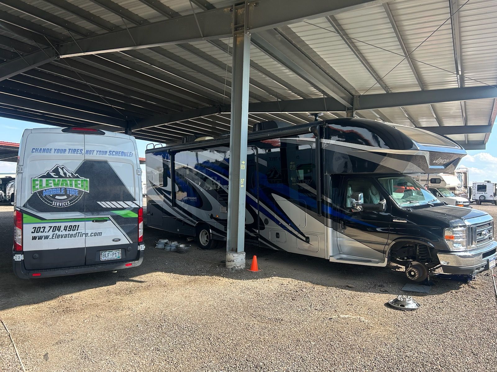 Two vehicles parked under a metal canopy: a black RV and a white van with logo, gravel ground.