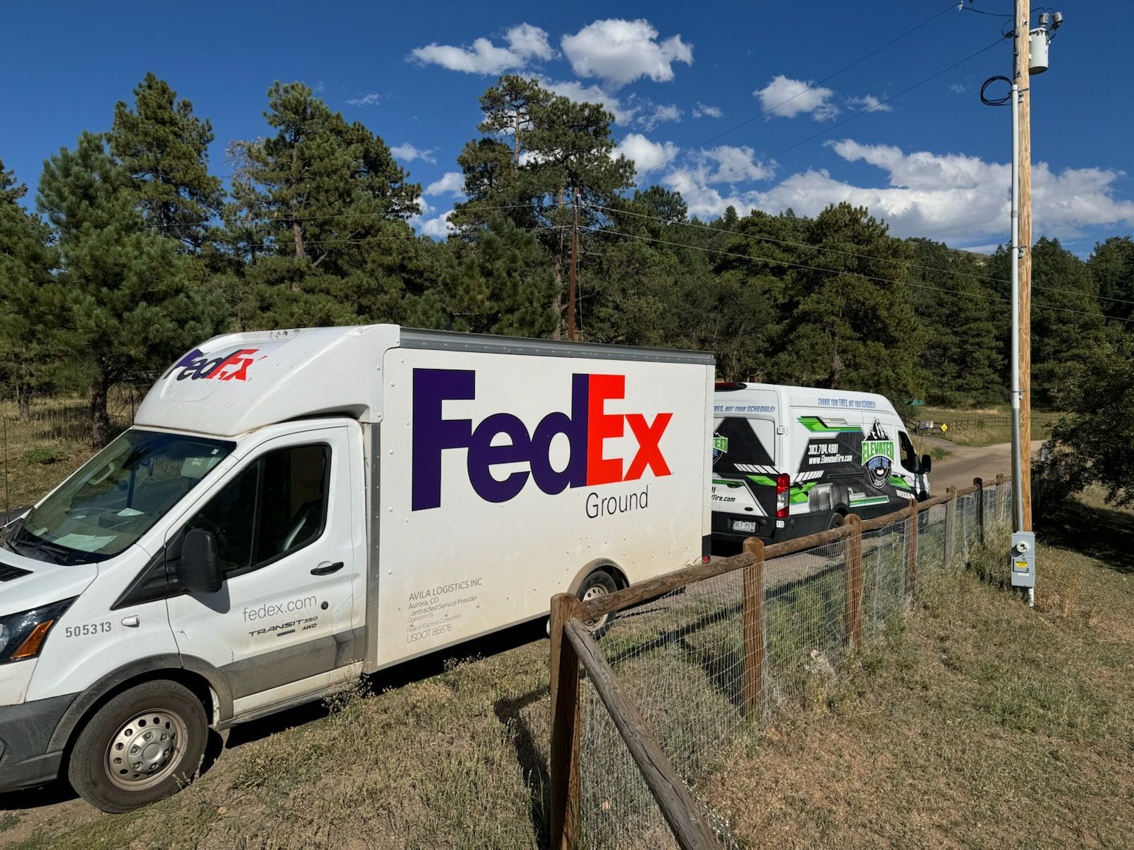 Two FedEx Ground vans on a dirt road next to a wooden fence with trees and a utility pole in the background.
