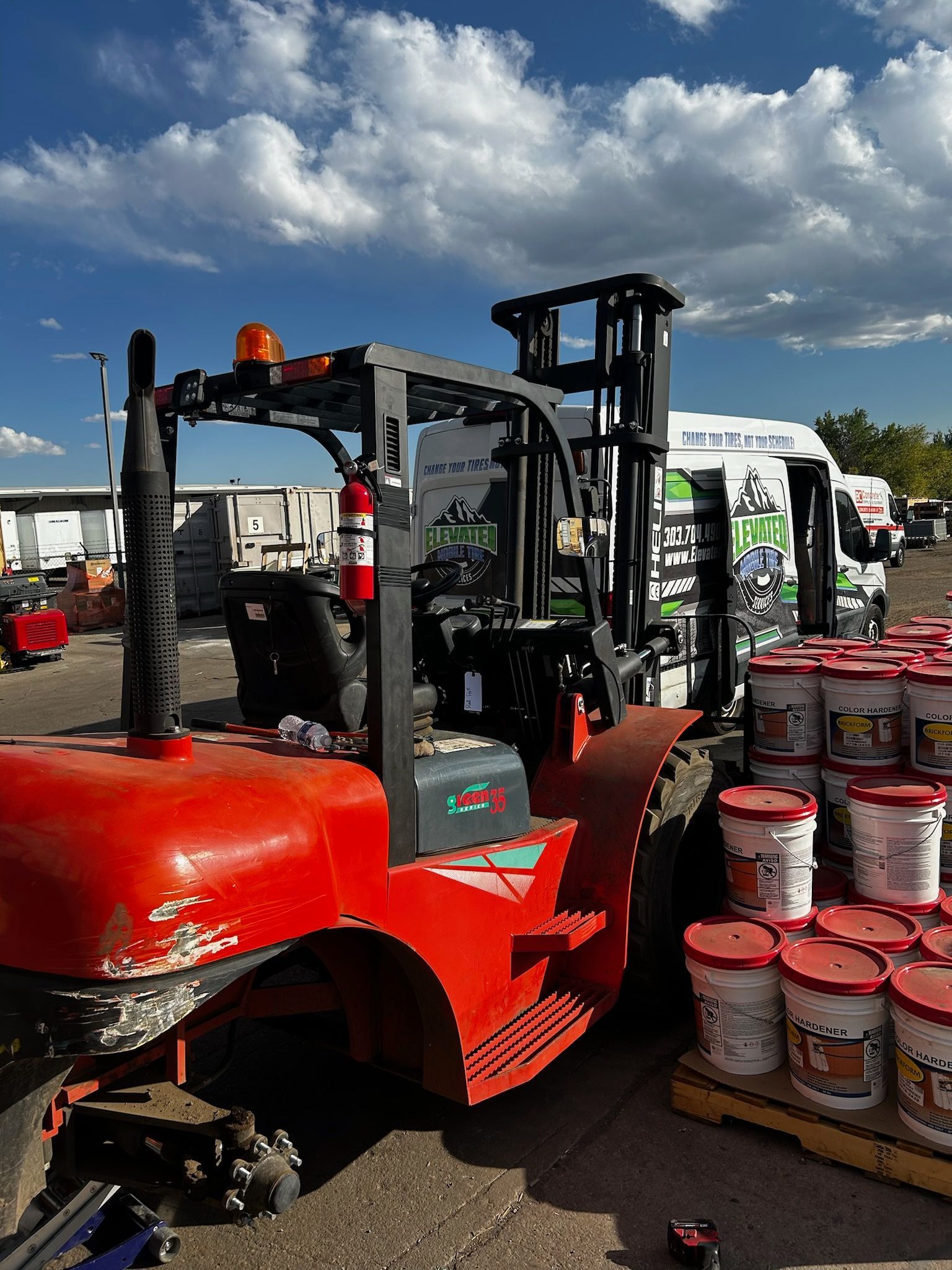 Red forklift in an outdoor setting with blue sky and clouds, next to pallets of paint buckets.