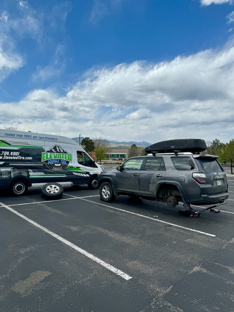 A gray SUV with a flat tire being towed by a tow truck in a parking lot under a blue sky.