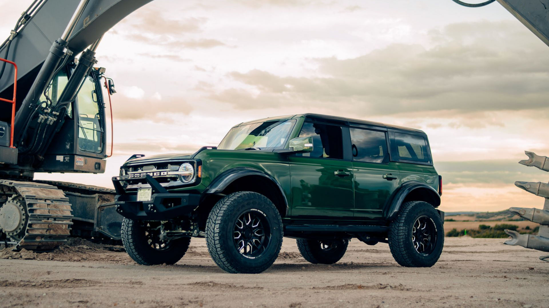Green Ford Bronco off-road vehicle with black accents, parked near construction equipment, overcast sky.