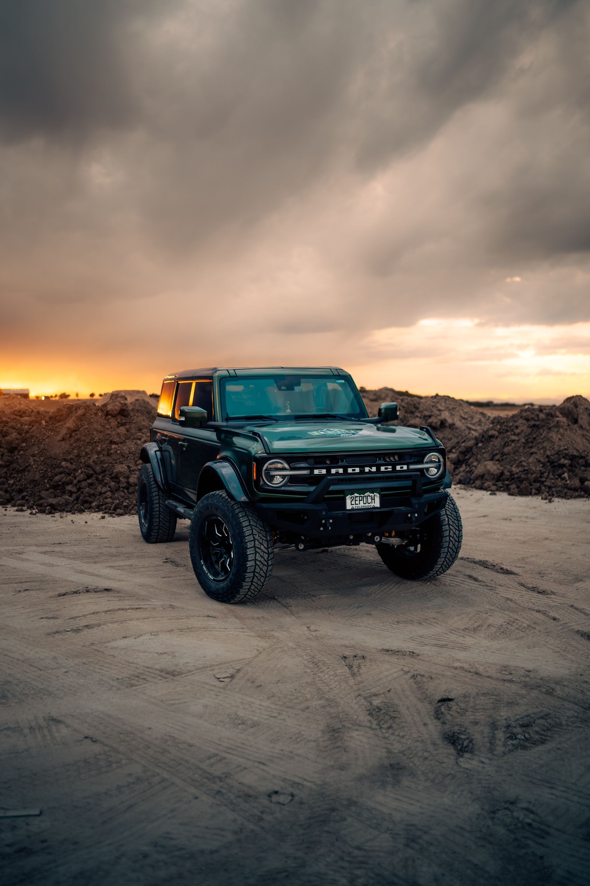 Dark green Ford Bronco off-road vehicle parked on sand, dramatic sunset backdrop.