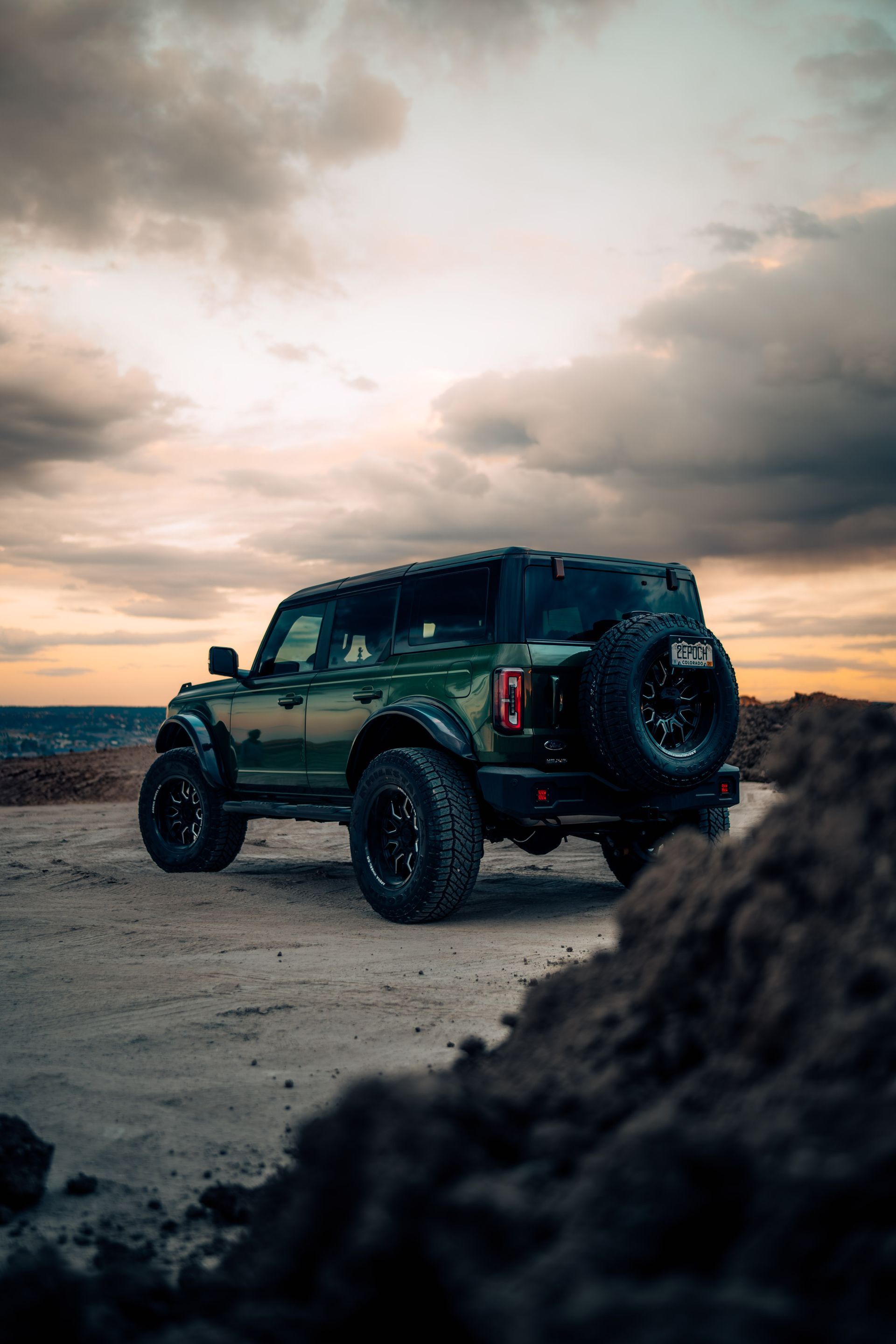 Green Ford Bronco SUV parked on a dirt road, silhouetted against a sunset sky.