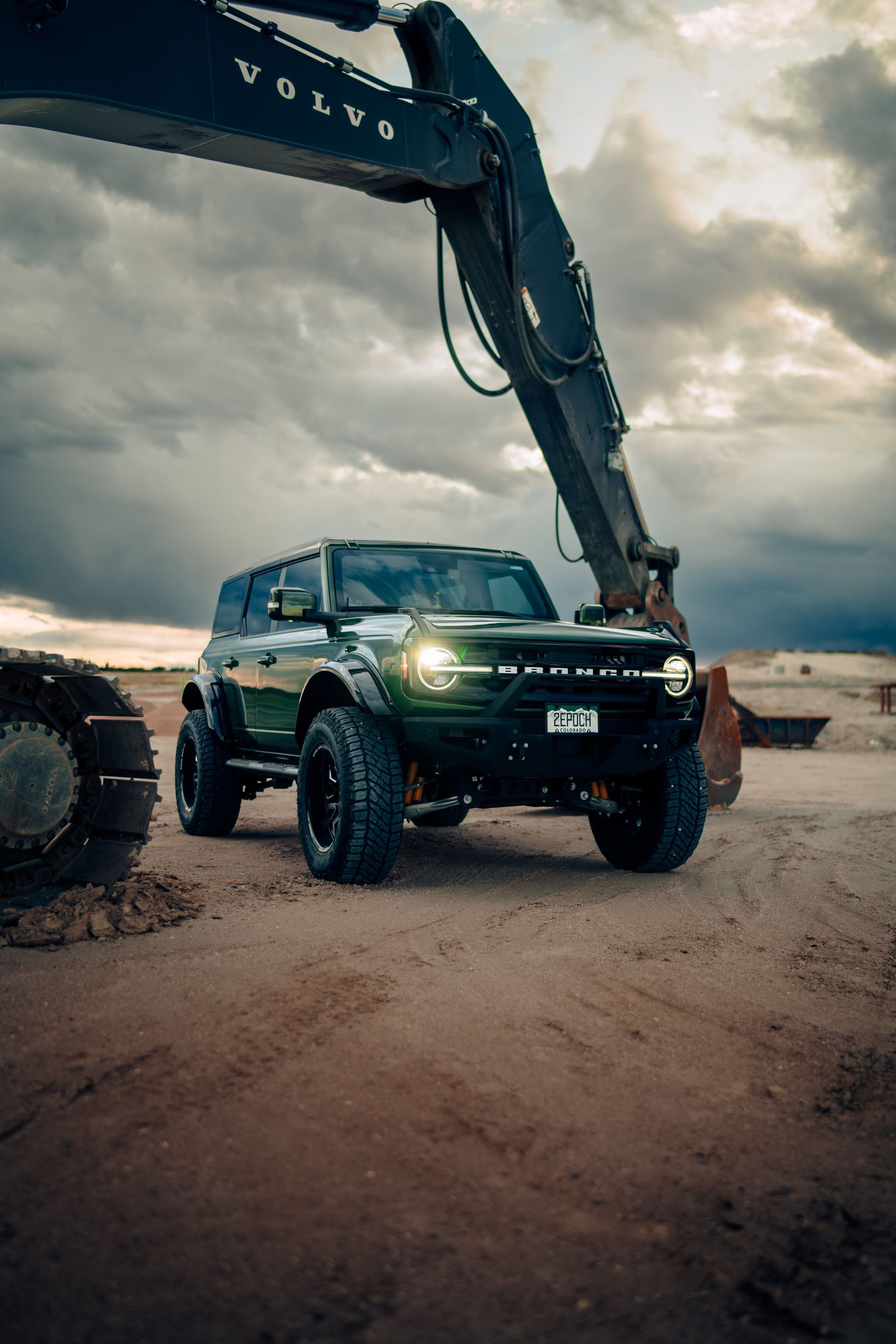 Green Ford Bronco parked in front of an excavator under a cloudy sky.