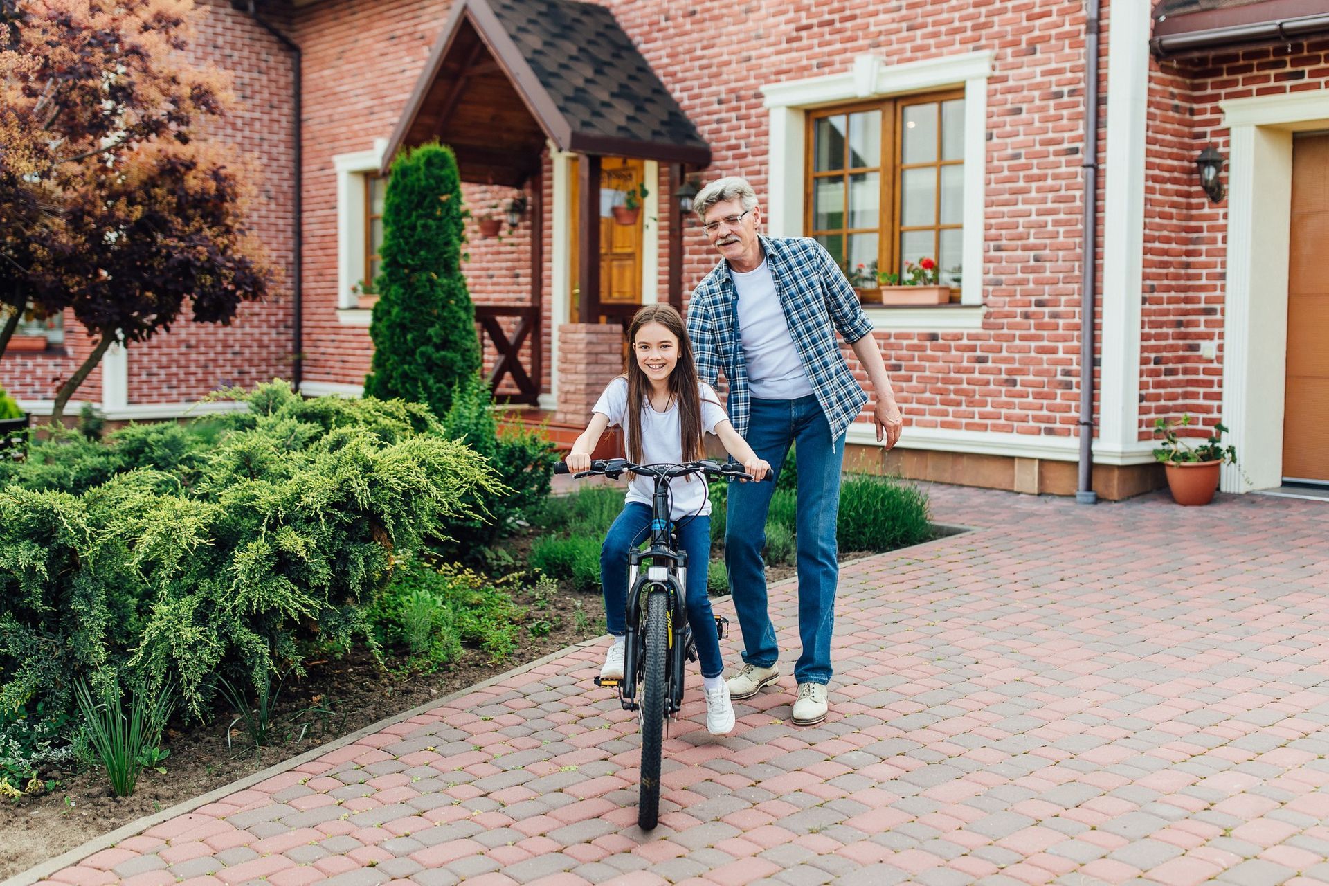 Boy teaching a girl how to ride a bike on a brick driveway in front of a brick house.