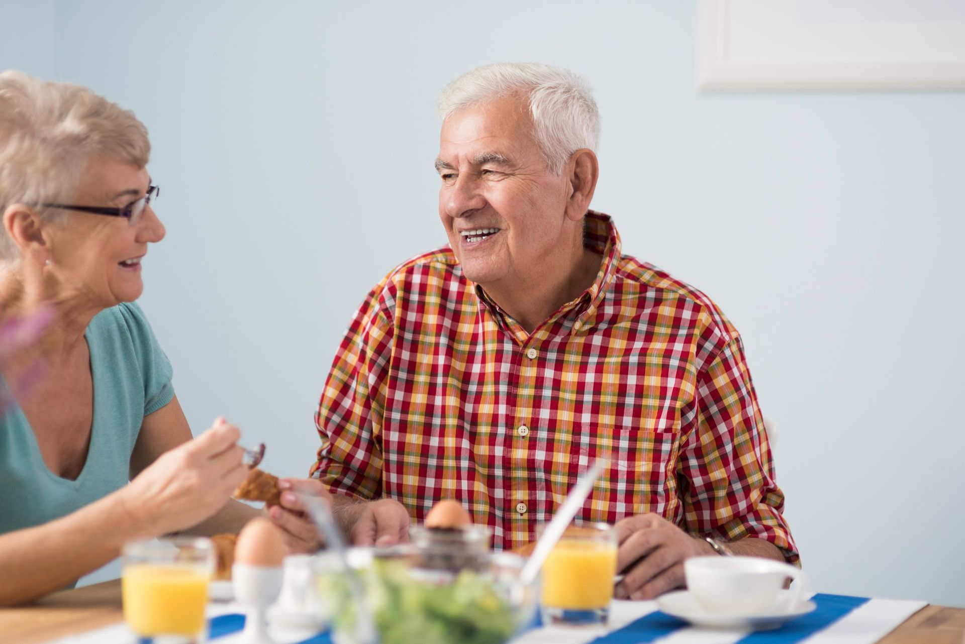 Senior couple smiling at each other at a breakfast table; orange juice, eggs, and salad present.