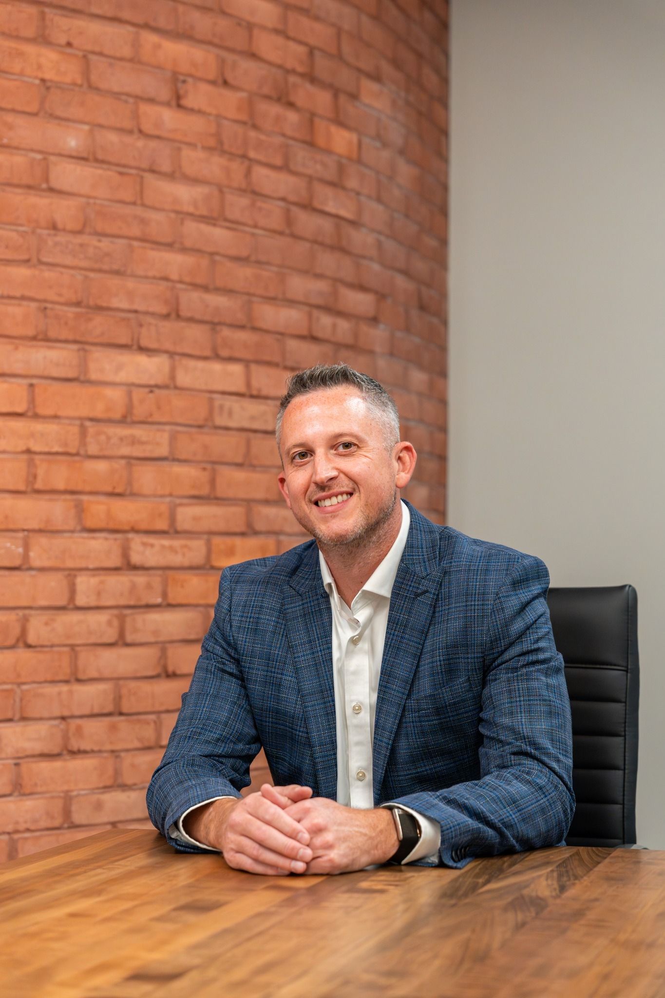 Man in blue patterned blazer at a wooden desk, arms crossed, smiling, brick wall background.