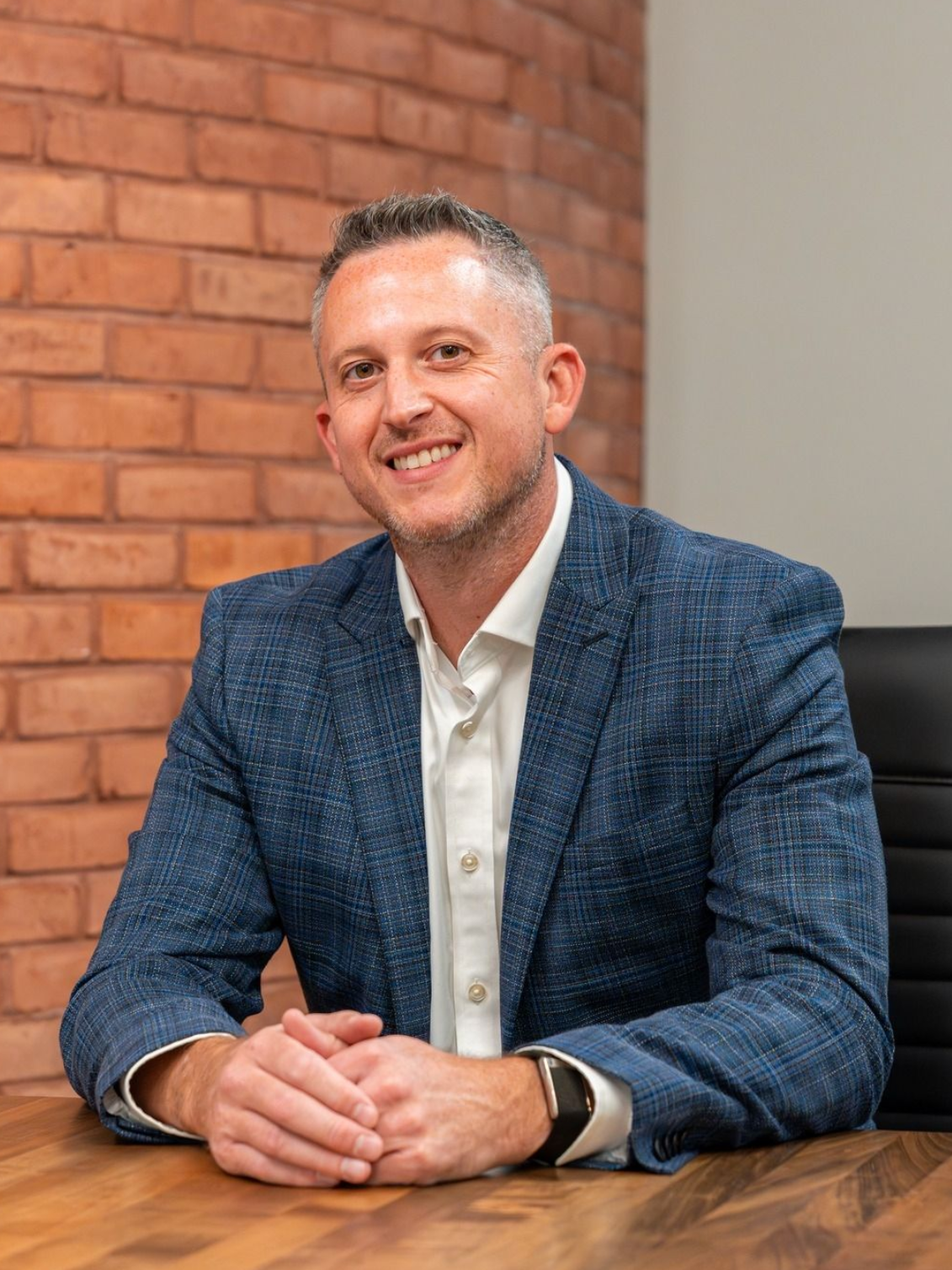 Man in blue blazer smiles at a wooden desk, brick wall background.