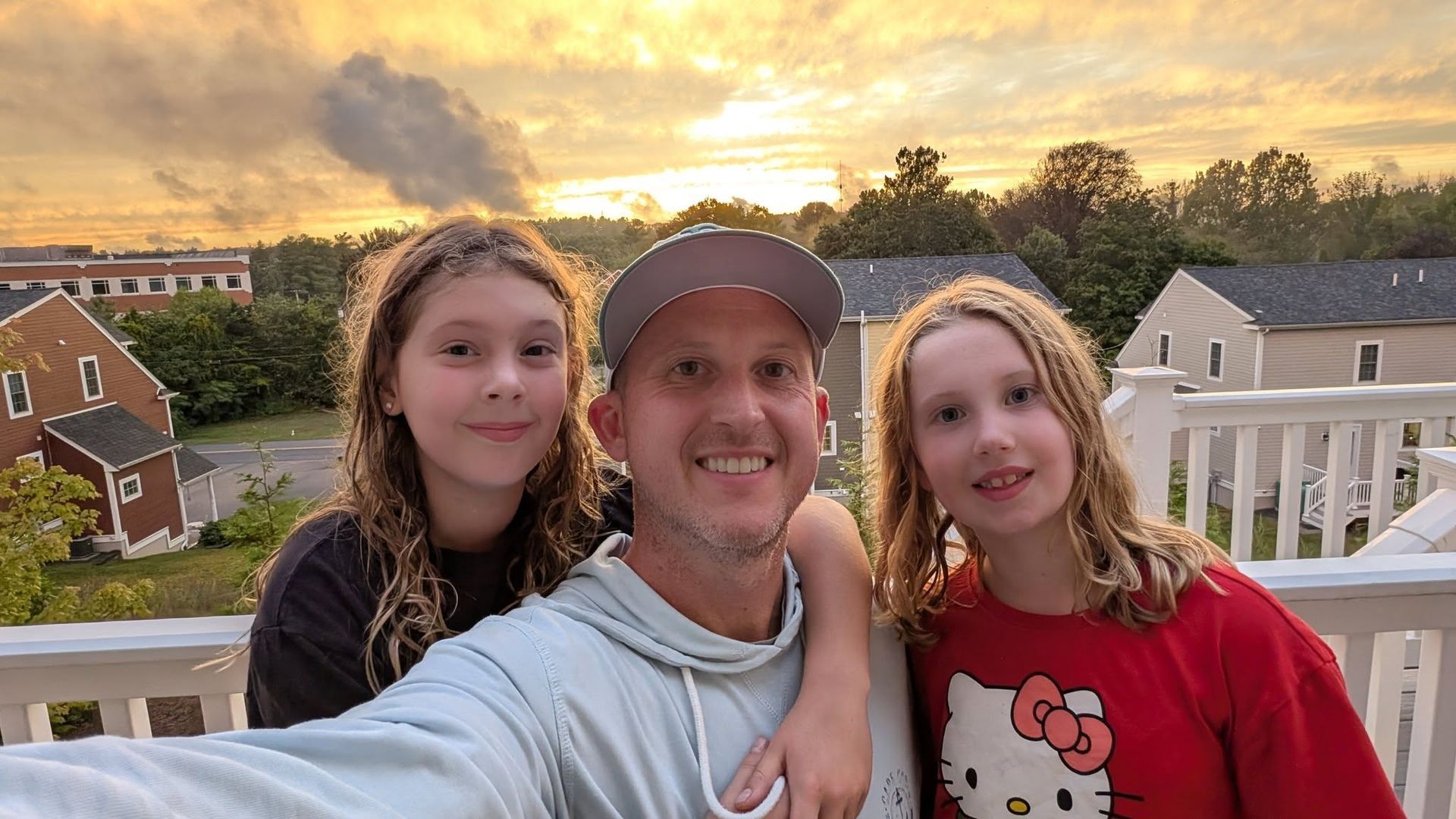Man smiles, selfie with two girls on a deck at sunset. Buildings in the background.