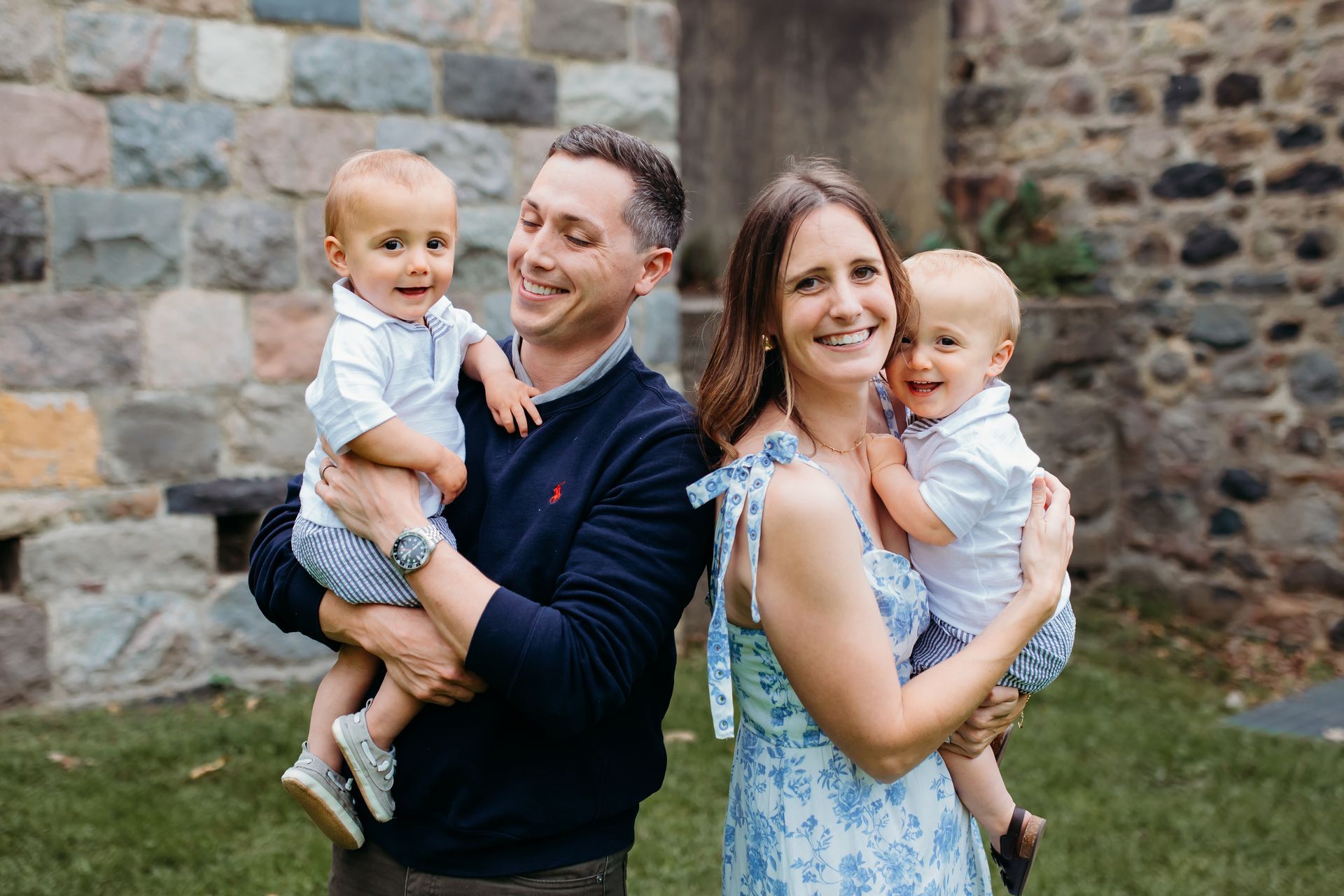Family of four poses outside: parents holding twin toddlers, all smiling.