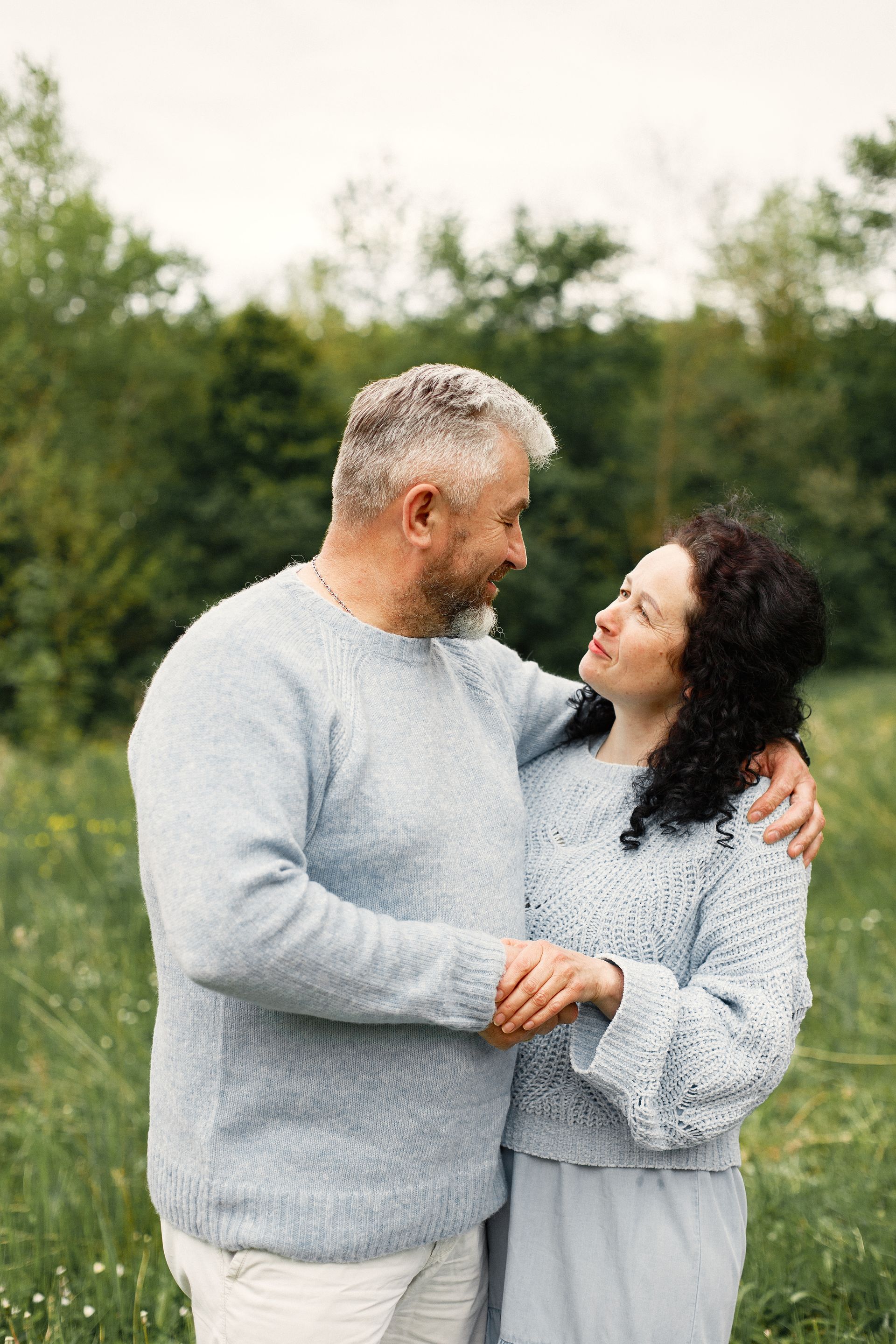 Couple in matching blue sweaters embracing in a grassy field, looking at each other.