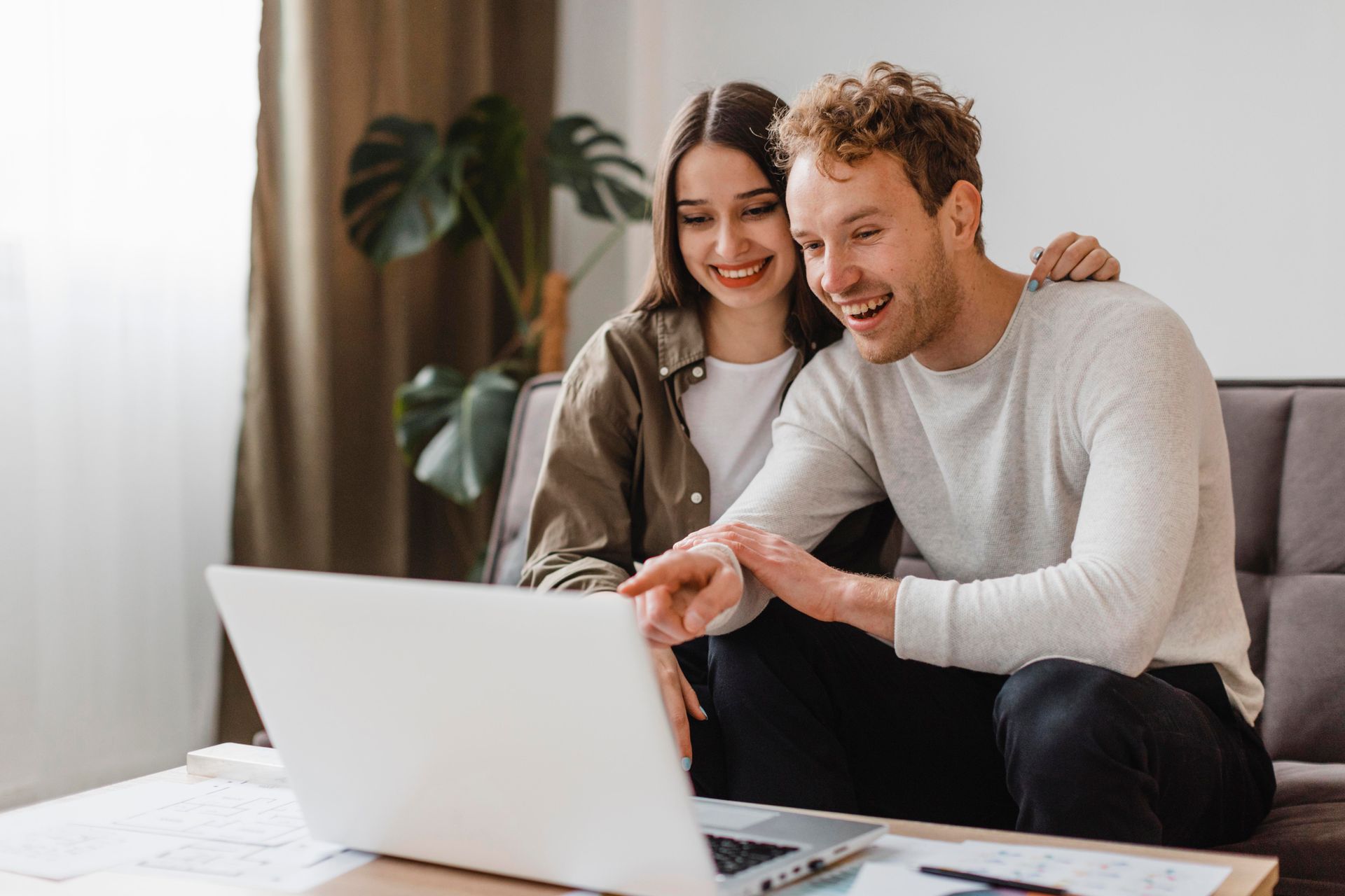 A smiling couple looks at a laptop, the man points to the screen, indoors with a plant.