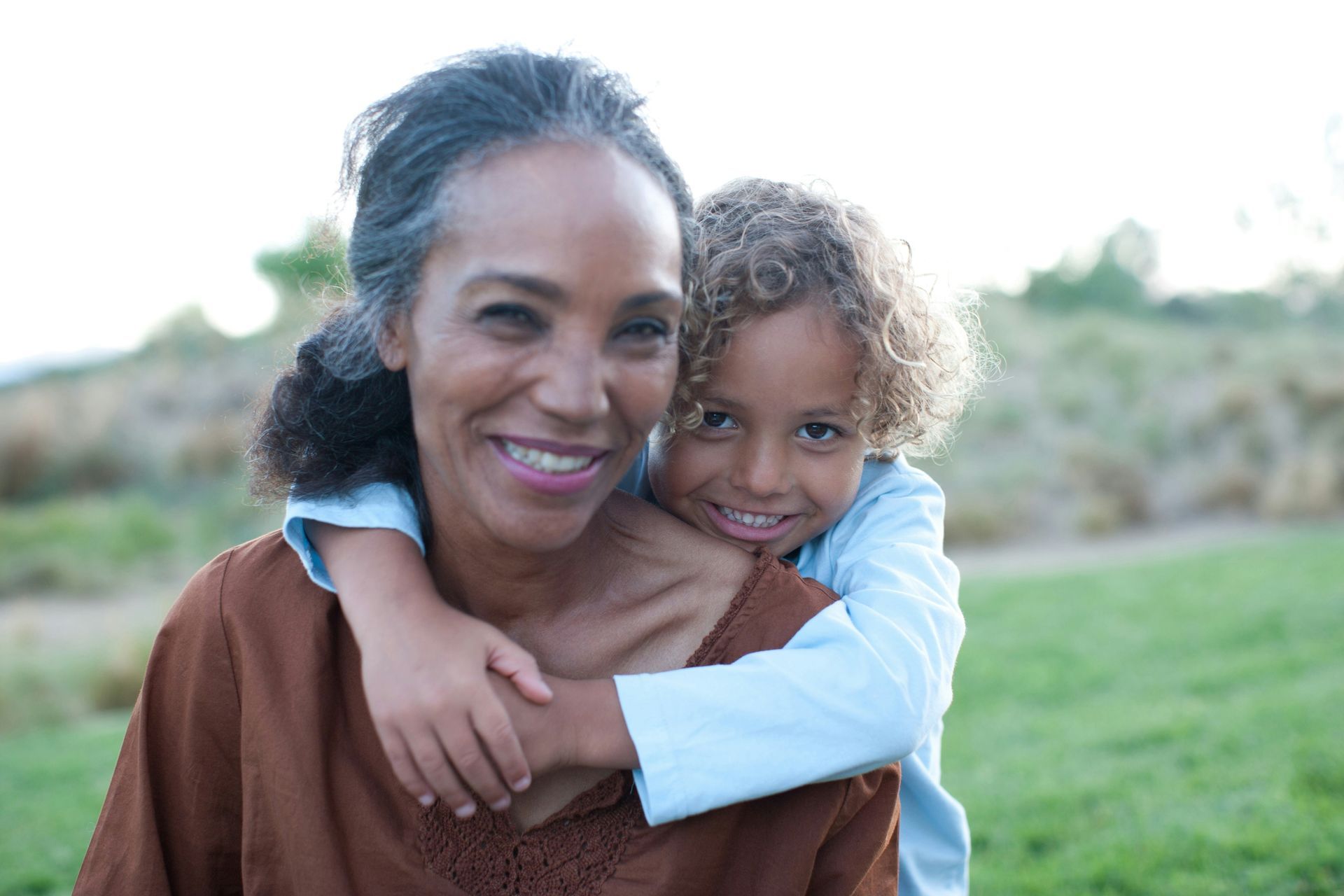 Grandmother smiling, embraces a young child, outdoors on a grassy field.