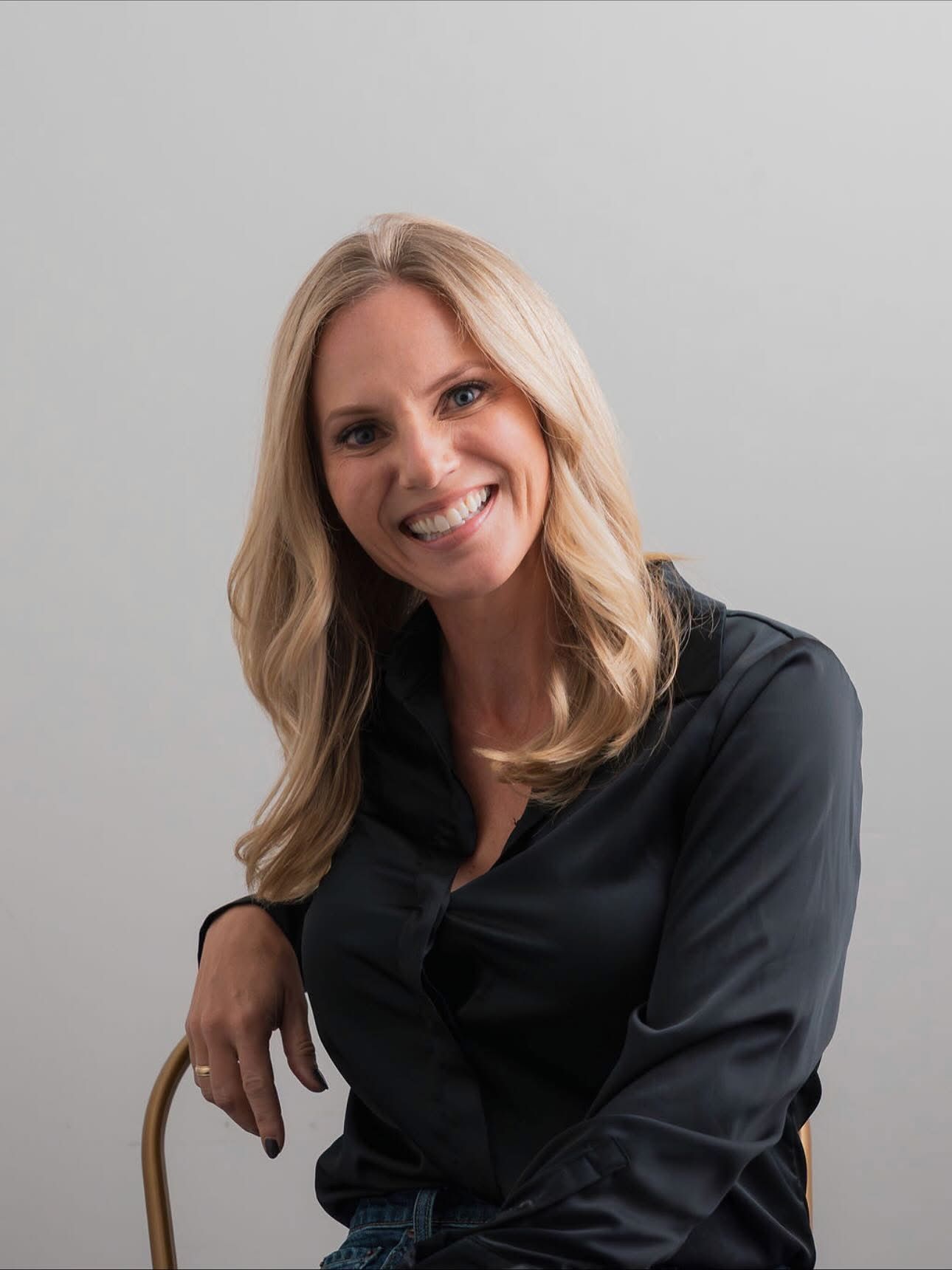 Blonde woman smiles, leaning on a chair, wearing a black satin shirt and jeans against a gray backdrop.